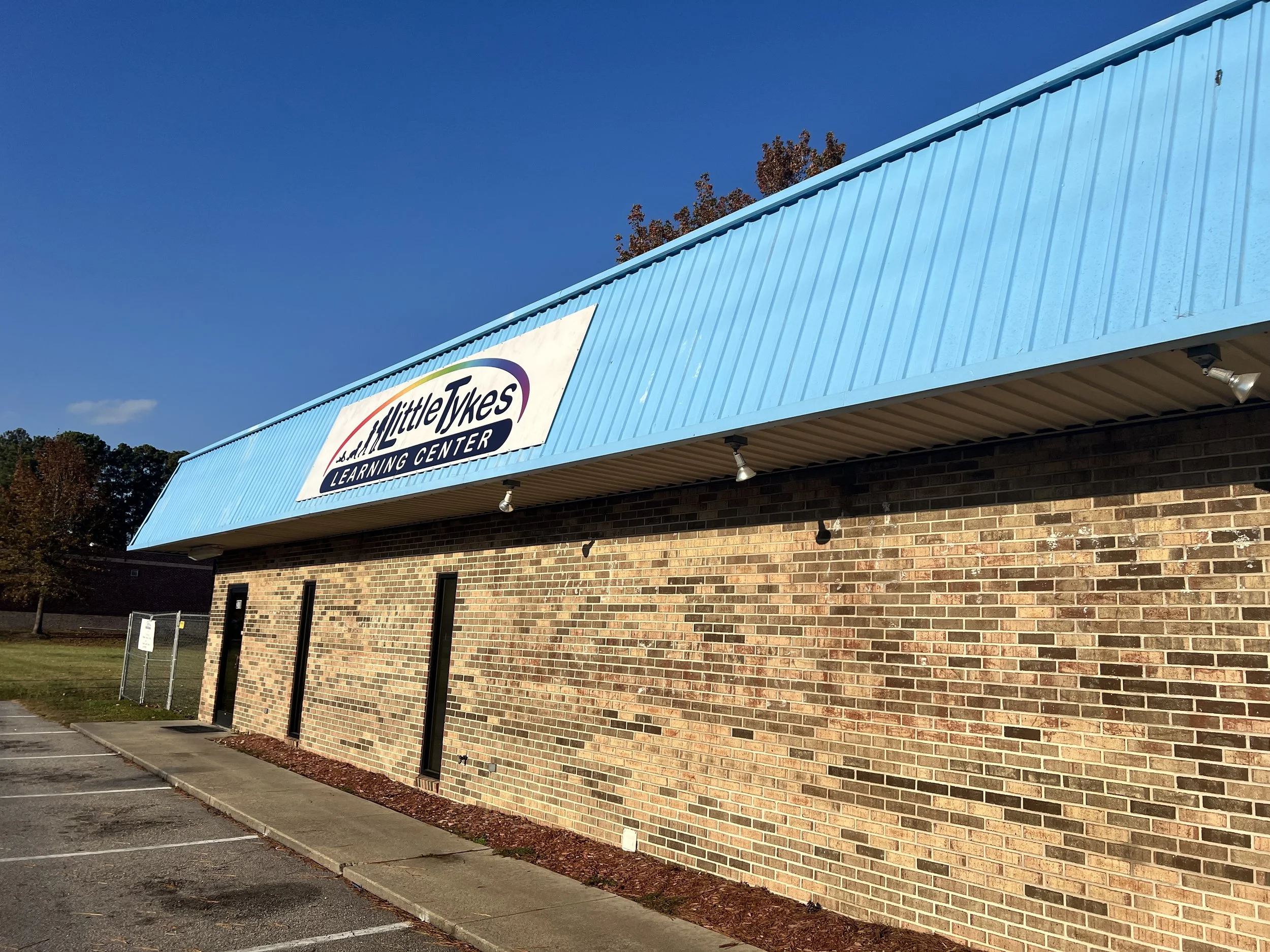Exterior view of a brick building with a blue metal awning displaying a sign that reads "Little Tykes Learning Center." The sky is clear with a few clouds, and there are trees with autumn-colored leaves in the background.
