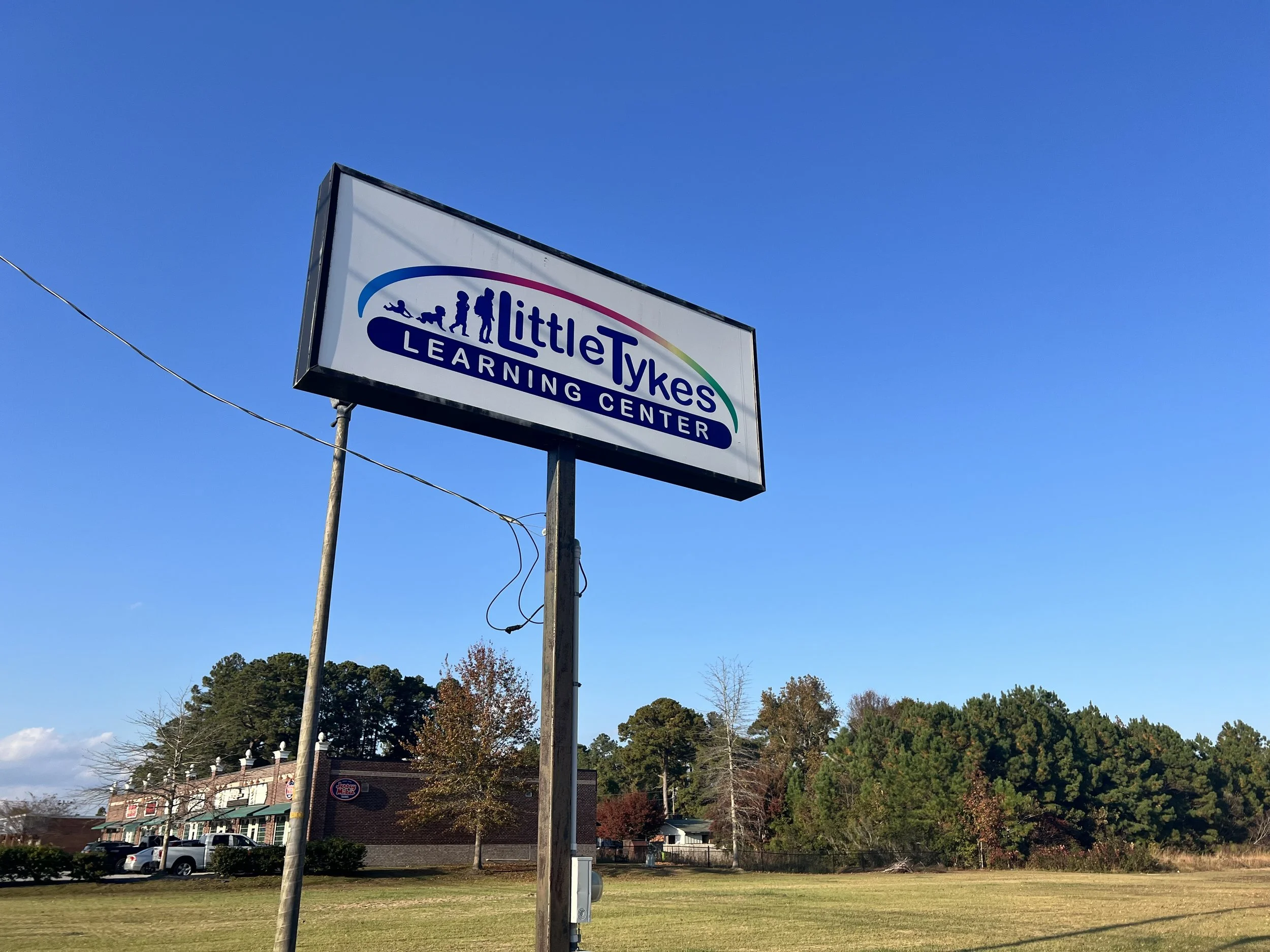 A sign for Little Tykes Learning Center on a tall pole against a clear blue sky. The sign has a rainbow-colored arc and silhouettes of children playing, with the text 'Little Tykes Learning Center.' In the background, there are trees, a parking lot with cars, and a building with a Burger King sign.