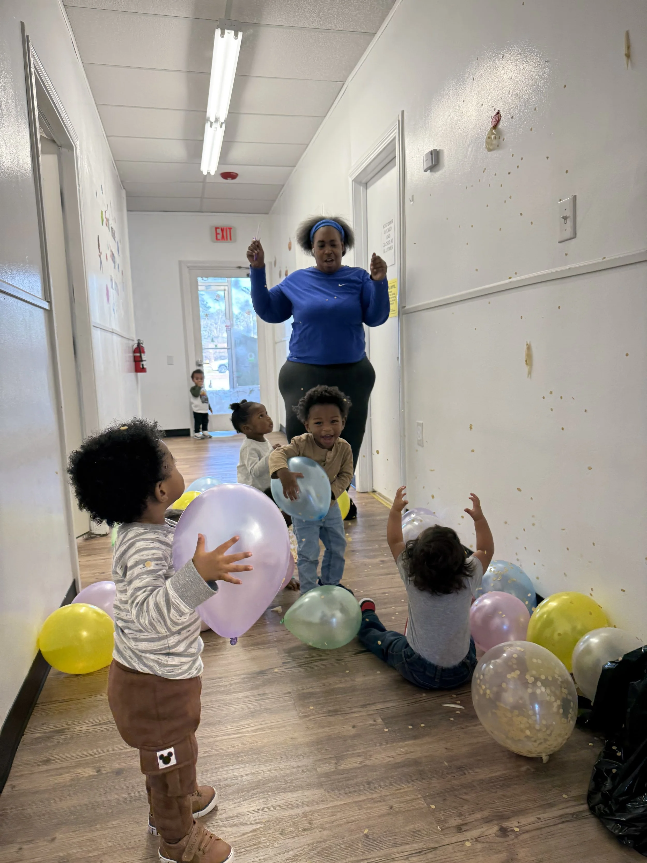 Children playing with balloons and confetti on the floor of a hallway, with a woman standing behind them and a glass door leading outside.