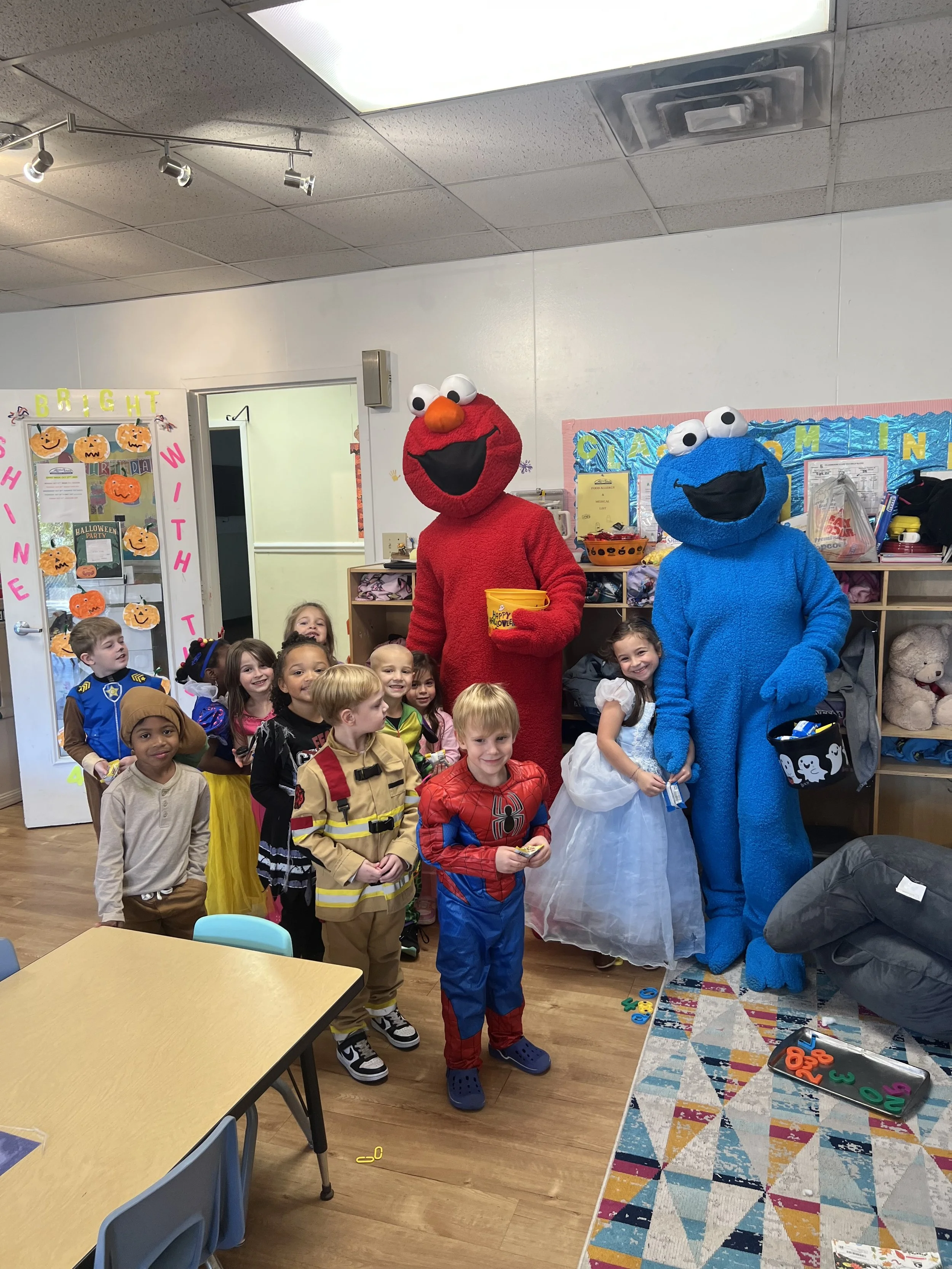 Children dressed in Halloween costumes standing and sitting around Elmo and Cookie Monster mascots in a classroom decorated for Halloween.
