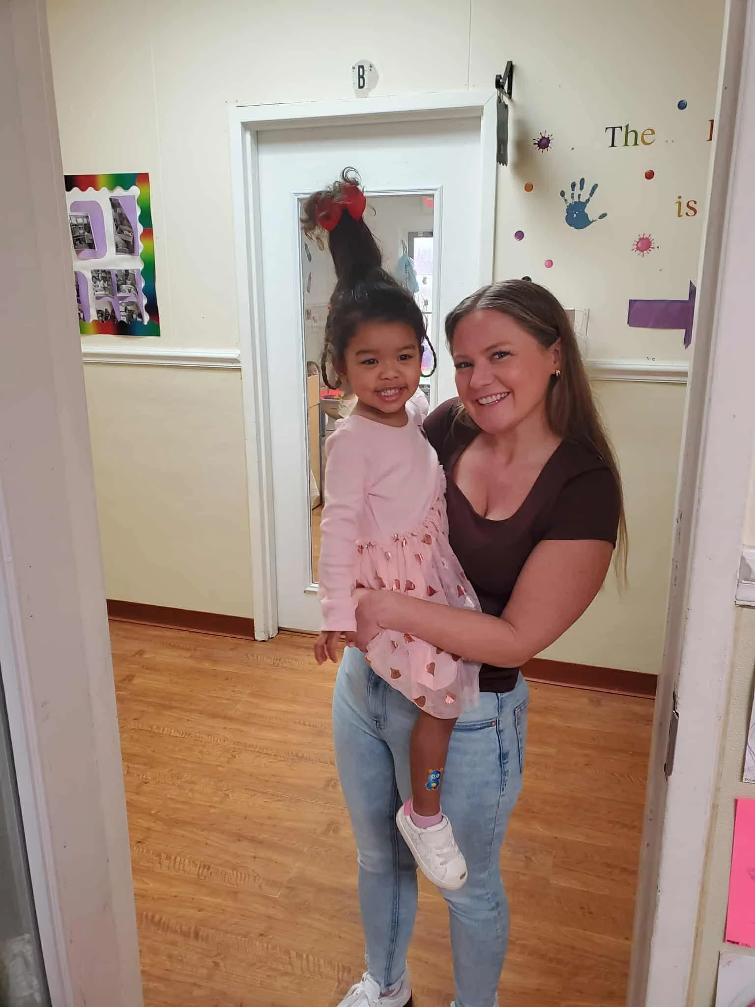 A woman with long hair, wearing a dark T-shirt and jeans, holding a smiling young girl with a pink dress and white sneakers, taking a mirror selfie in a room with light-colored walls and wooden flooring.