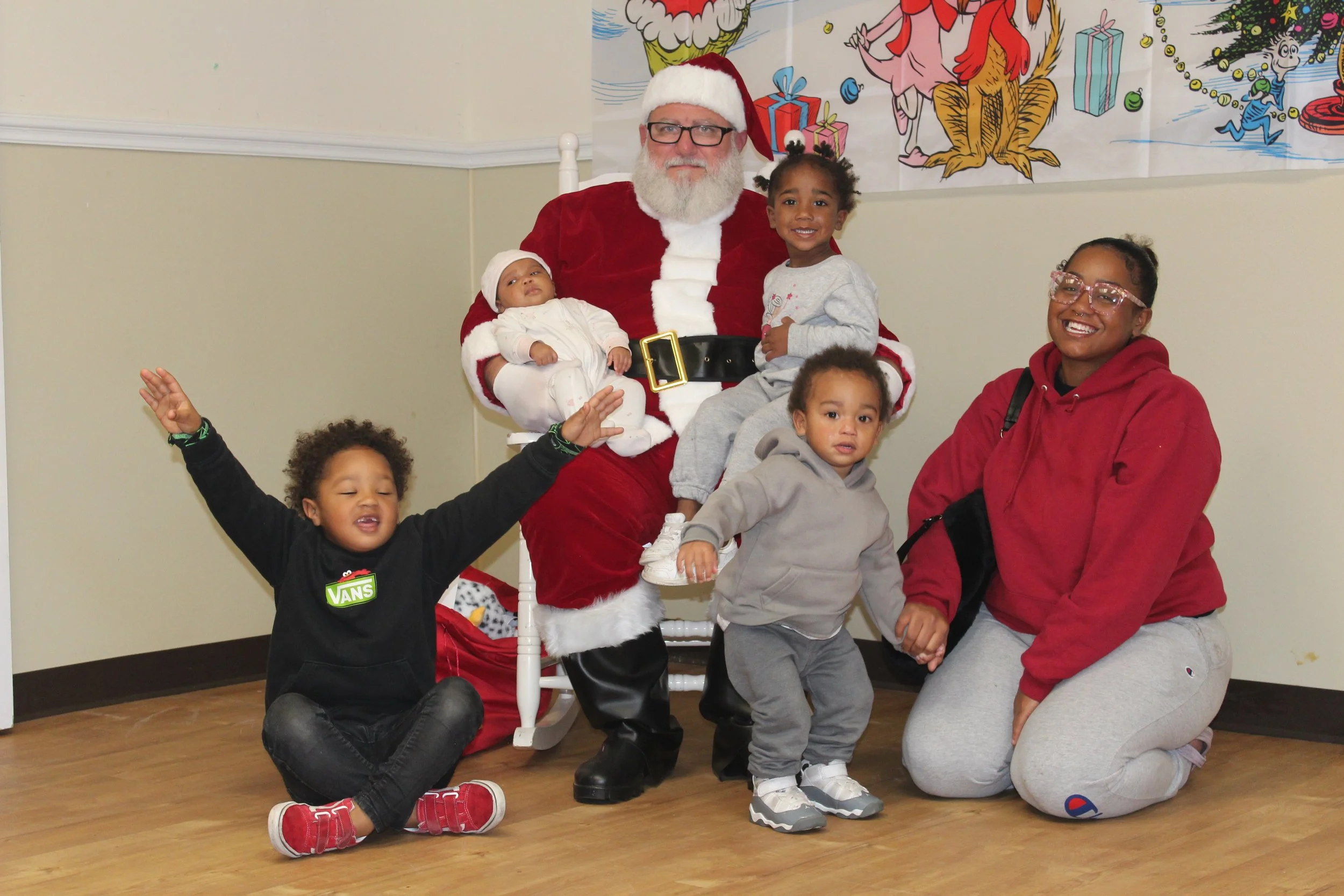 A man dressed as Santa Claus sitting on a white chair surrounded by children at a holiday event. The children are smiling and posing with Santa, with some kneeling or standing. A colorful festive backdrop featuring holiday-themed illustrations is in the background.