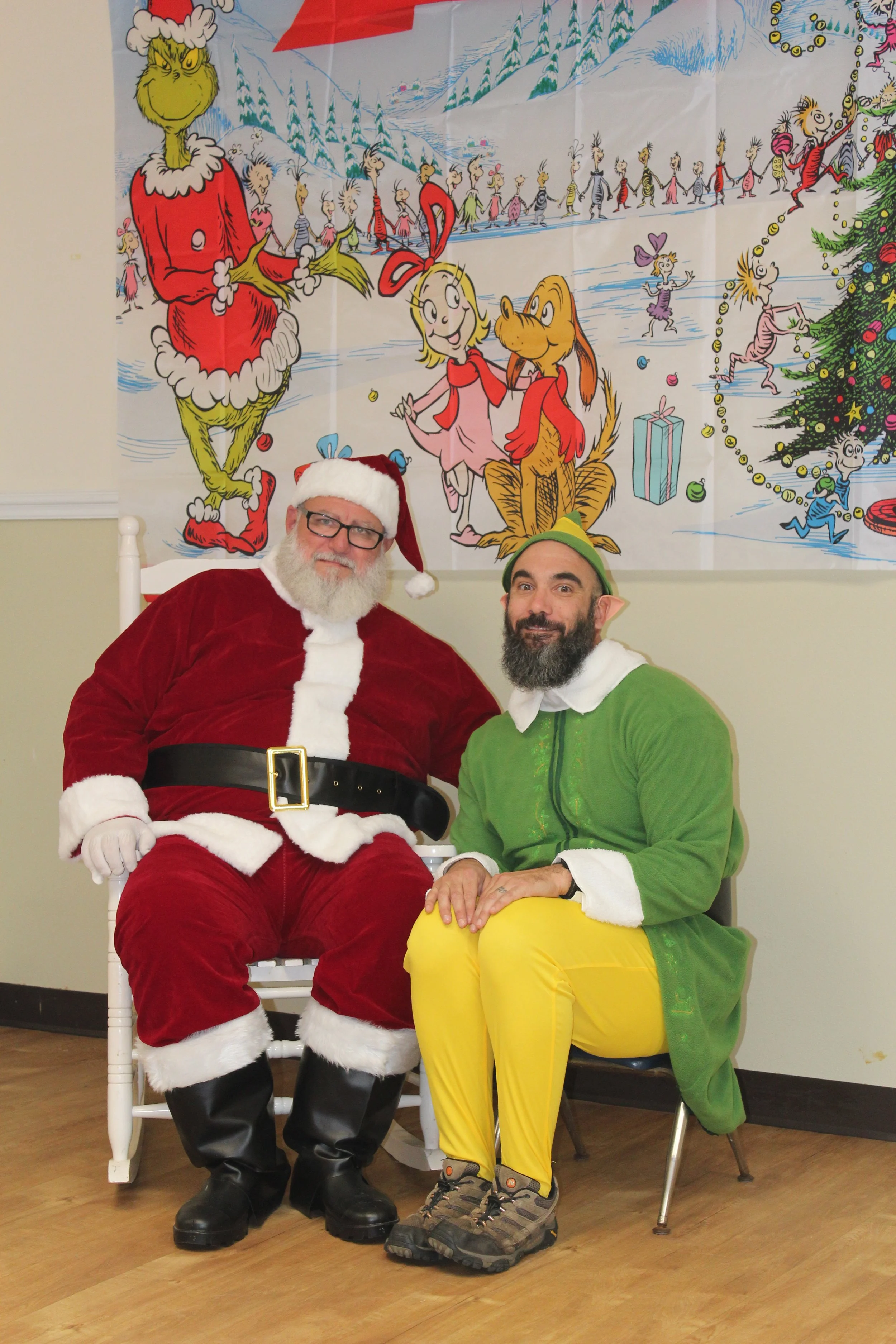 Two men dressed as Santa Claus and an elf sitting indoors in front of a colorful Christmas-themed poster, smiling for the photo.