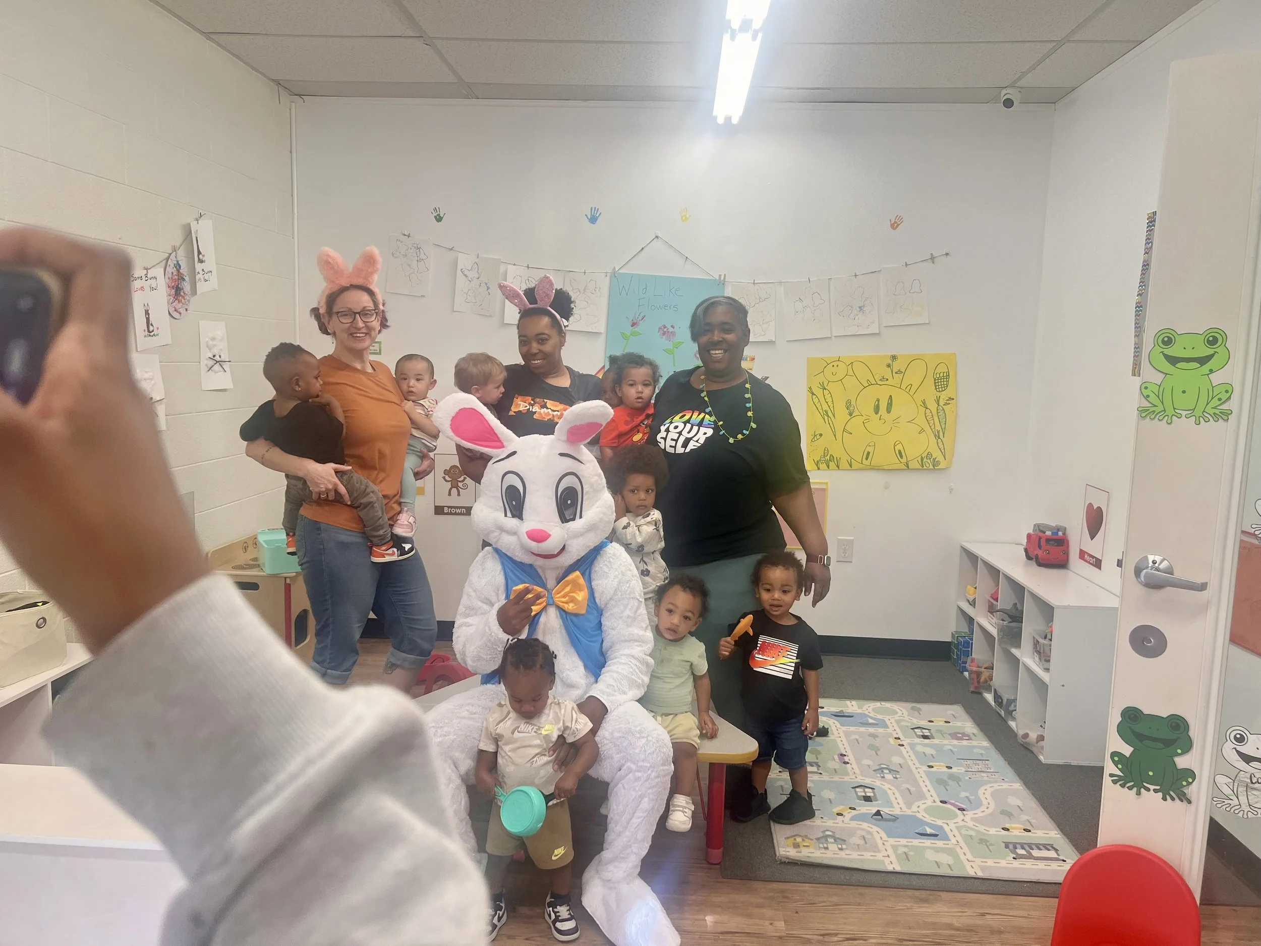 A group of children and three adults celebrating Easter in a classroom decorated with bunny-themed artwork. One adult dressed as the Easter Bunny is seated with children around her, and two women wearing bunny ears stand among the kids. Some children are holding toys or coloring books.