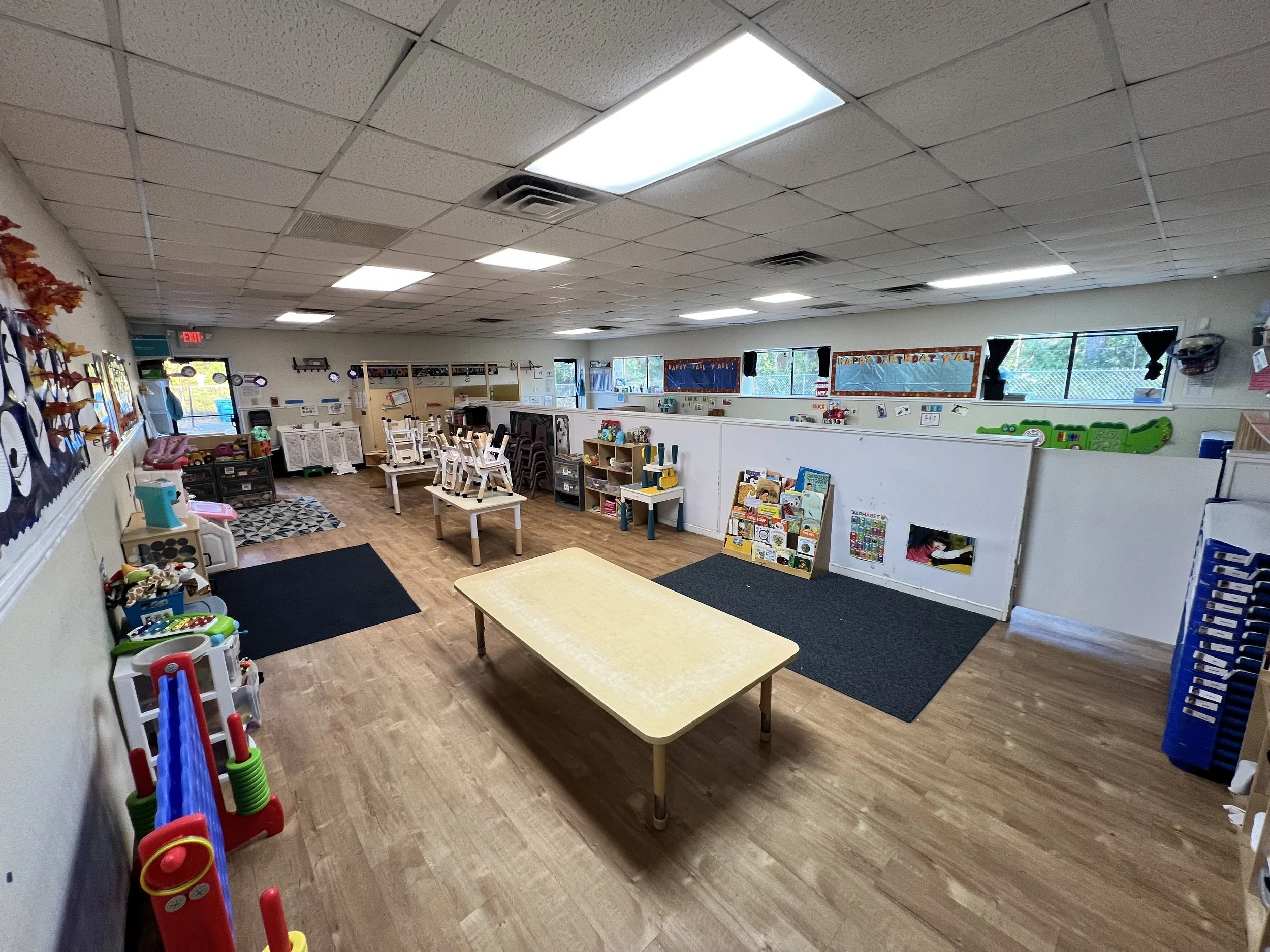 A classroom with tables, bookshelves, and educational toys, decorated for fall with orange and red leaves on the walls and windows, and a sign wishing a happy birthday on the wall.