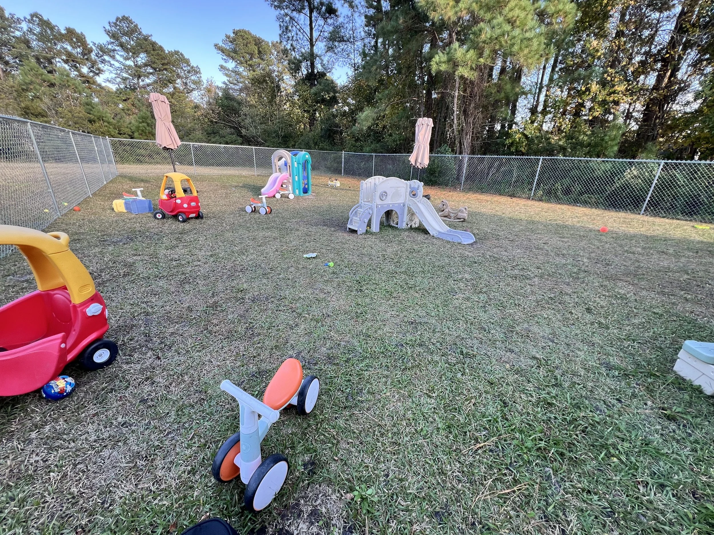 Children's outdoor play area with small slide, ride-on toy cars, and fencing surrounded by trees.