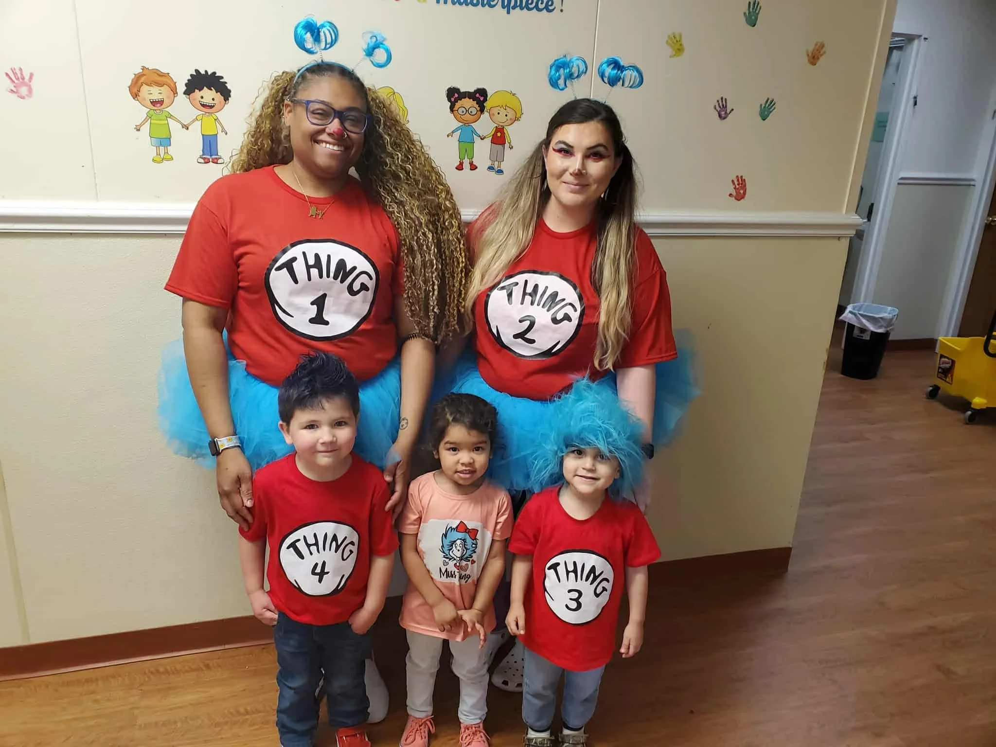 Two women and four children posing indoors at a celebration, with a wall decorated with colorful handprints and cartoon children illustrations in the background. The women are wearing red shirts with the words 'Thing 1', 'Thing 2', 'Thing 3', and 'Thing 4' written inside white circles.
