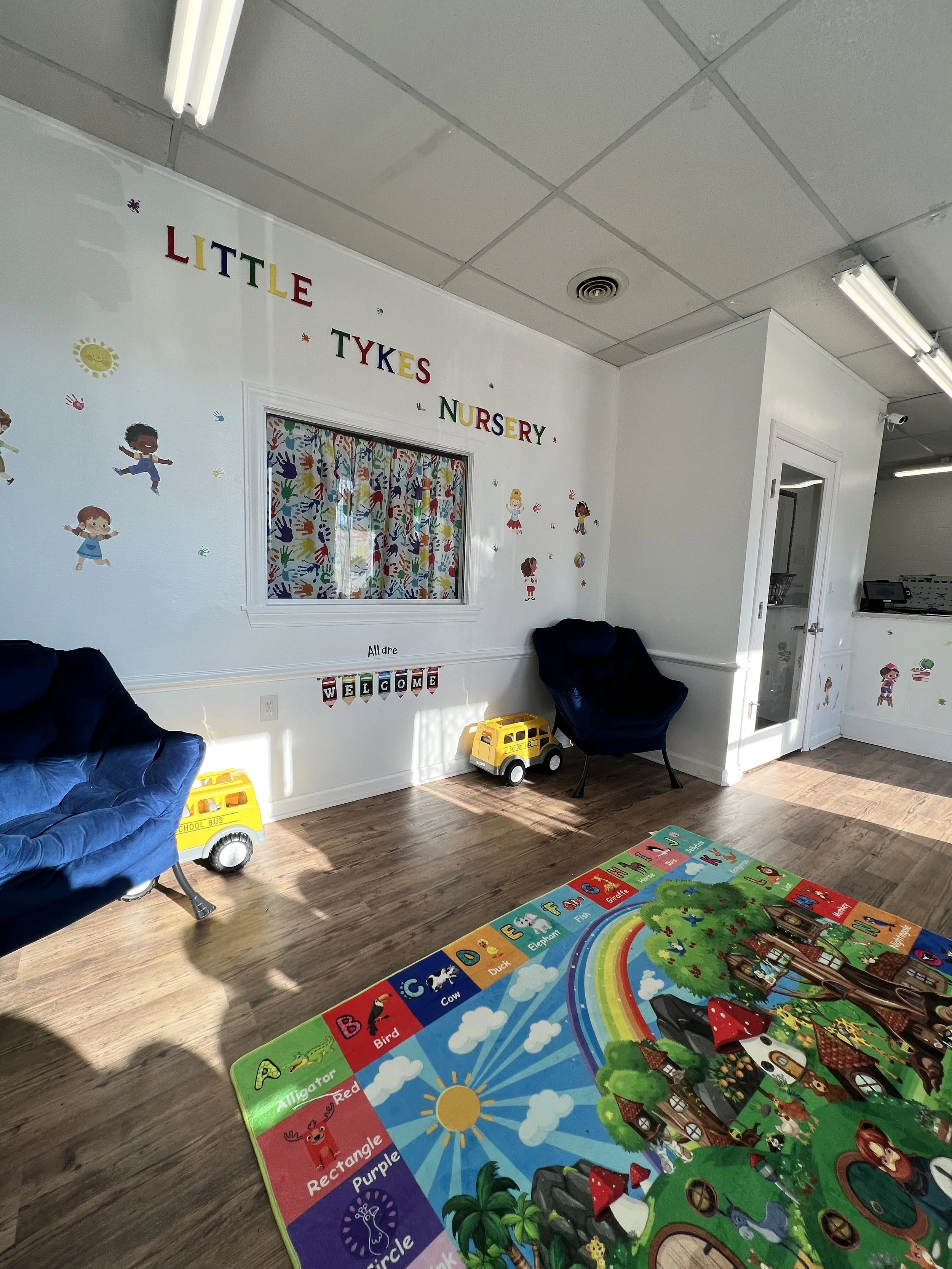 Interior of Little Tykes nursery featuring wall decorations of children and animals, a colorful alphabet floor mat with animal images, and small school bus toys on the floor.