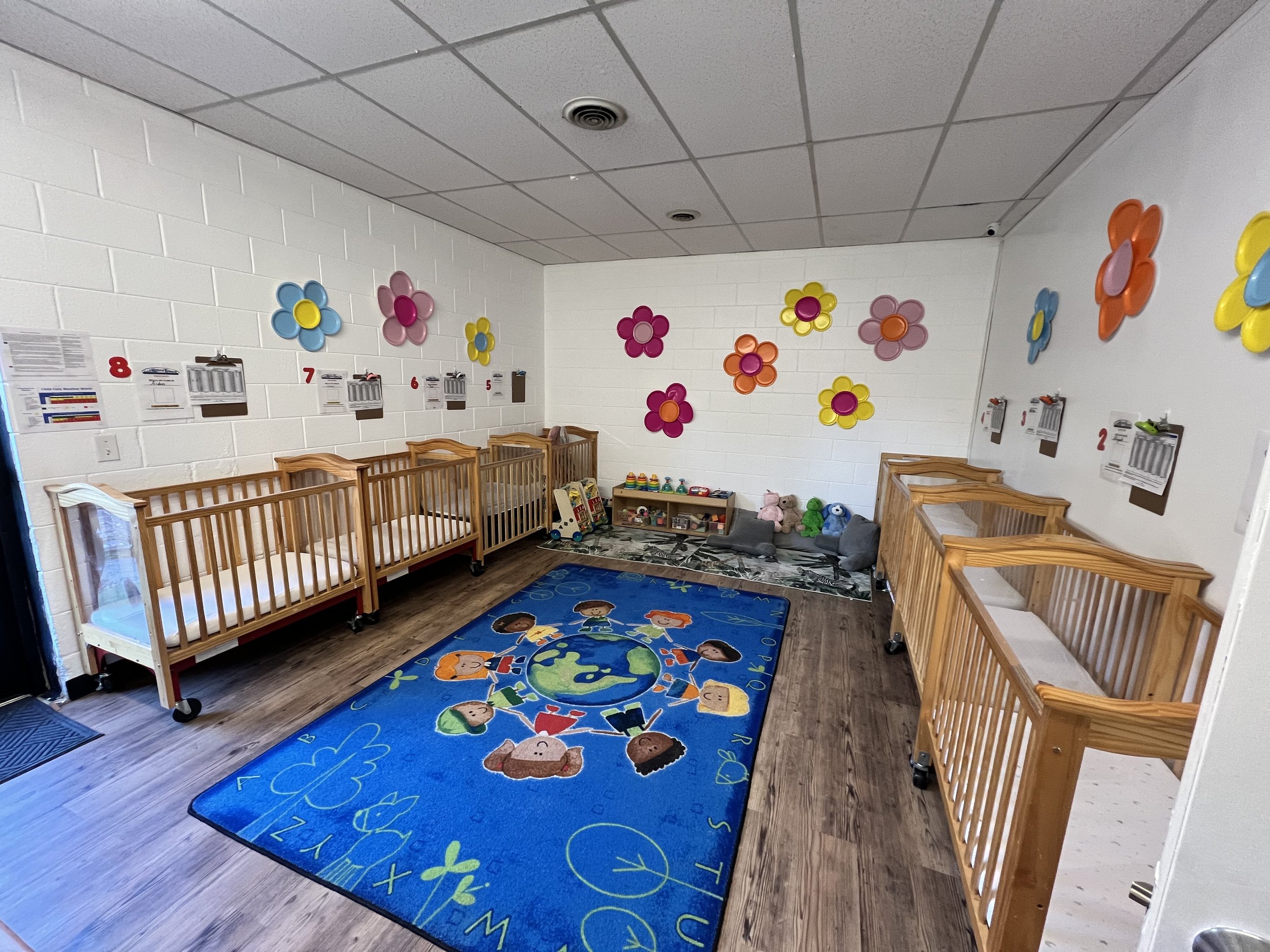 Childcare room with wooden cribs, colorful flower wall decorations, a blue rug featuring children and Earth illustrations, and toys along the wall.