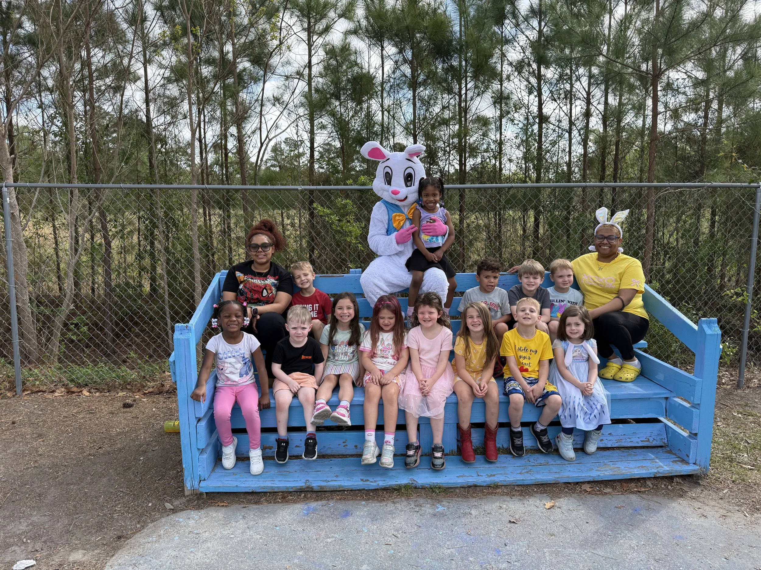 A group of children and two adults sitting on a blue platform in a wooded outdoor setting, with a person dressed as a white Easter Bunny character in the center.