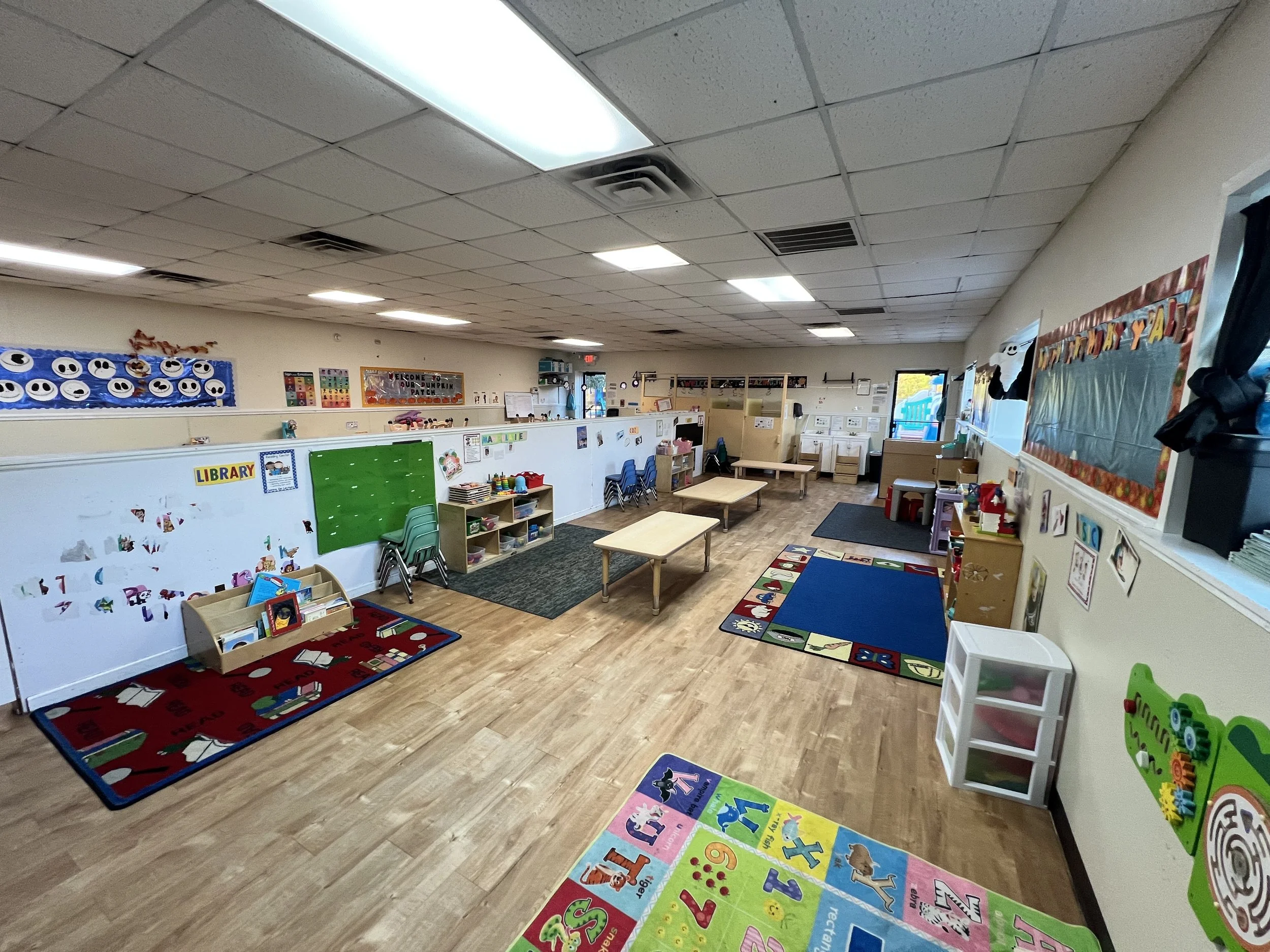 Interior of a preschool classroom with colorful educational decorations, small tables and chairs, a red and green alphabet rug, and storage shelves along the walls.