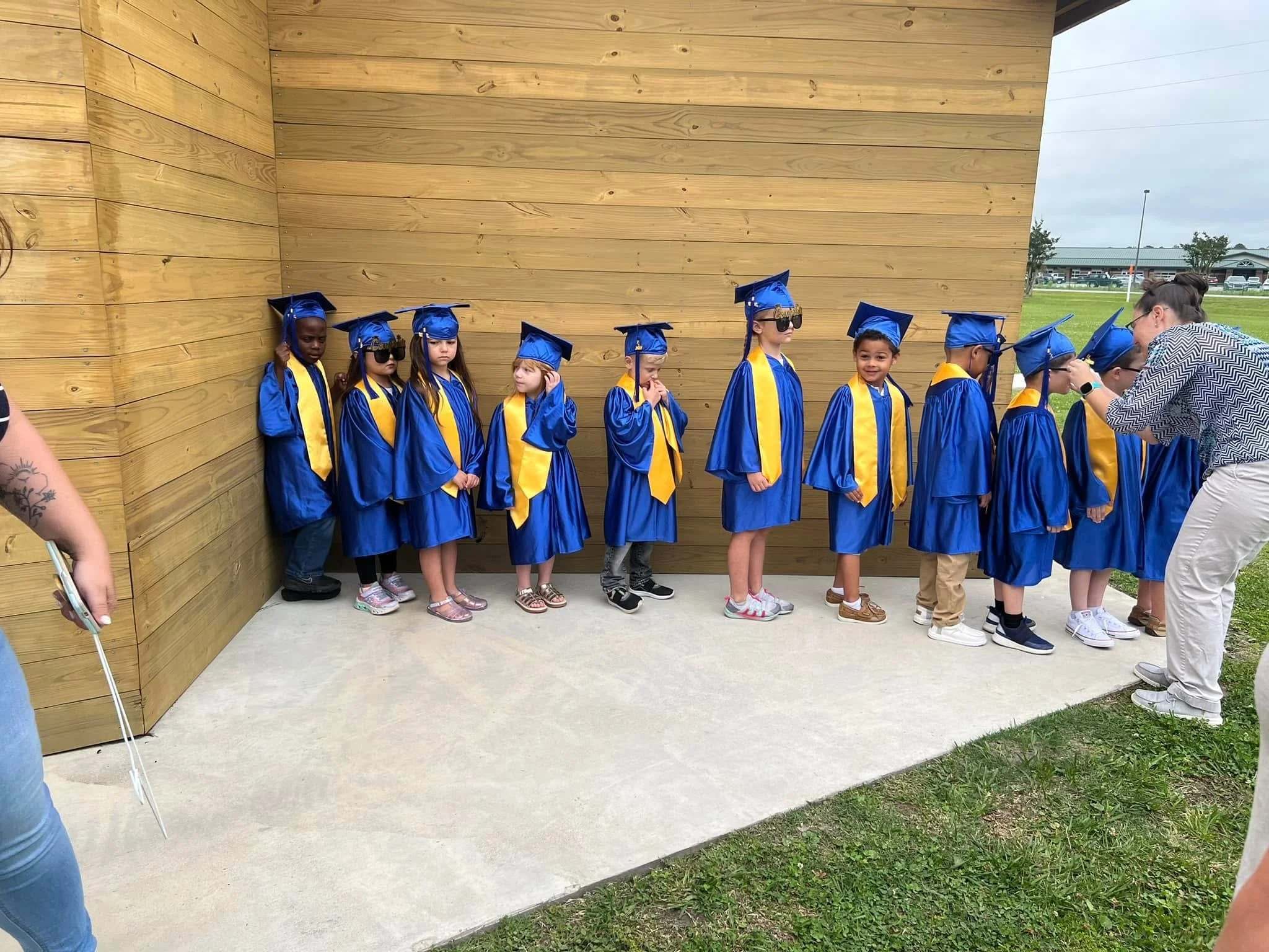 A group of children dressed in blue graduation gowns and caps, some wearing sunglasses, standing outside against a wooden wall, with a woman on the right enrolling the children in a graduation ceremony.