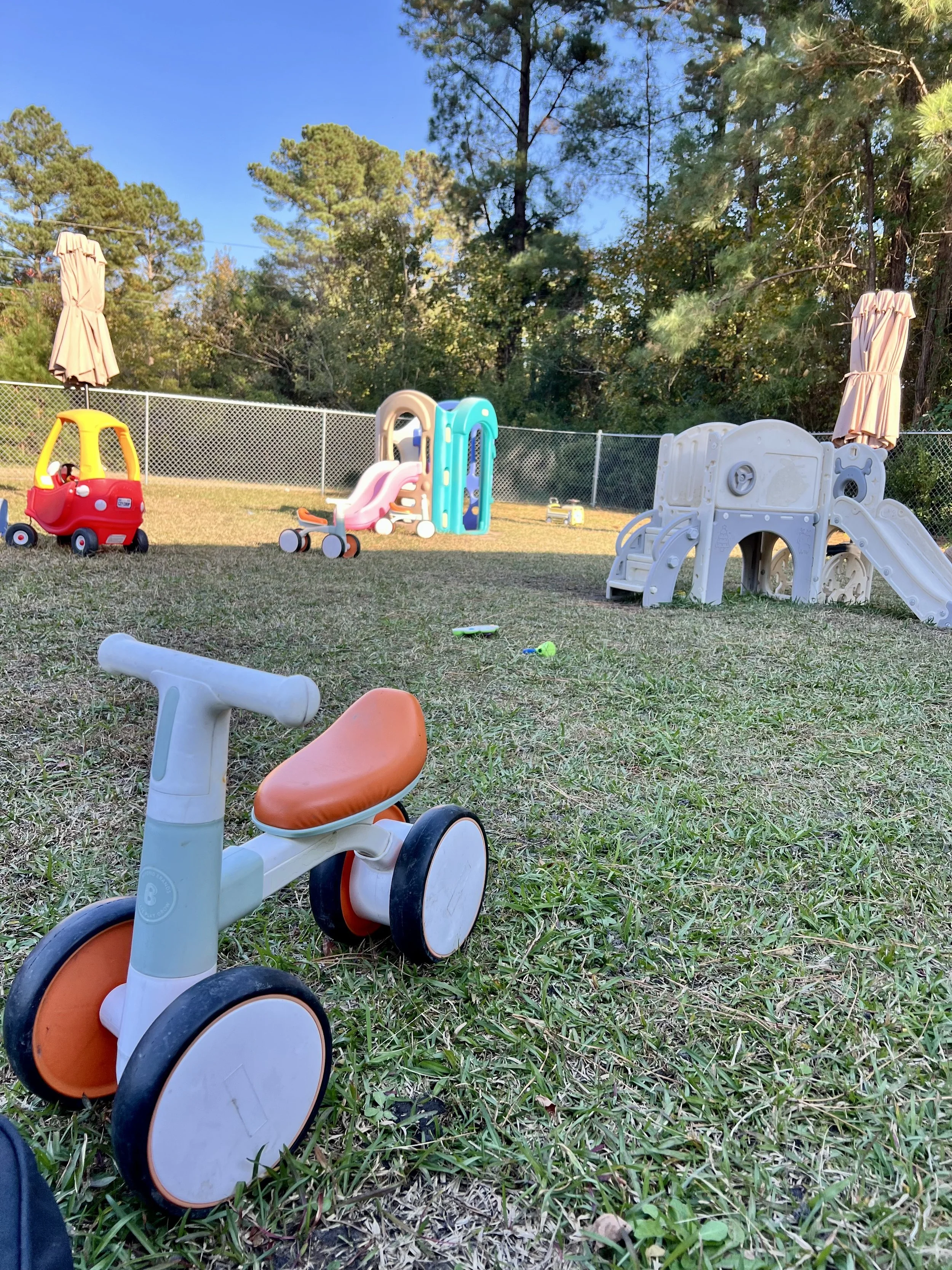 A fenced backyard with grass, trees, and playground equipment. There are two umbrellas, a small red and yellow toy car, a plastic slide, a climbing toy, small toys on the grass, and a white and orange tricycle in the foreground.