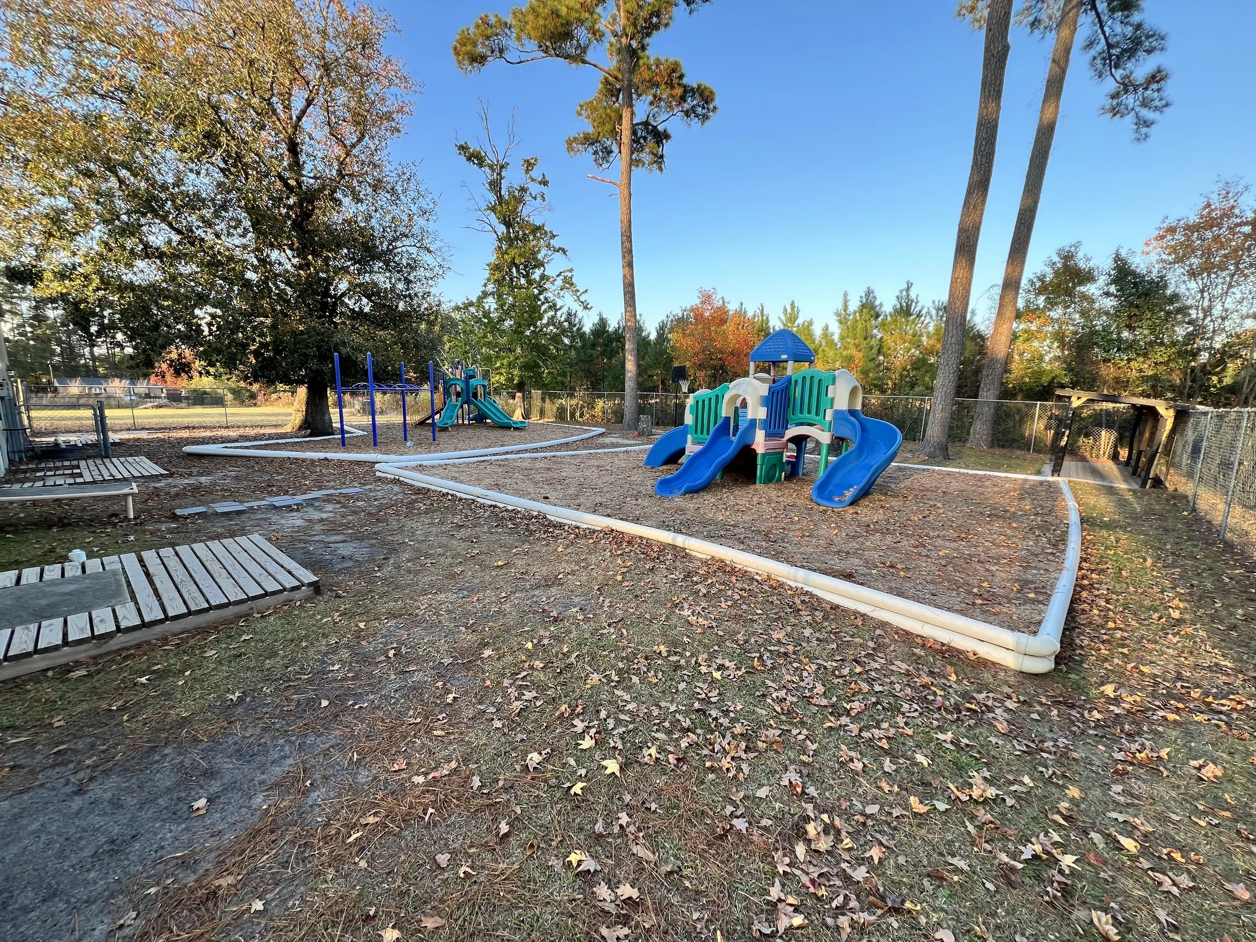 Children's playground with small slides, swings, and play structures, surrounded by trees with autumn leaves, under a clear blue sky.
