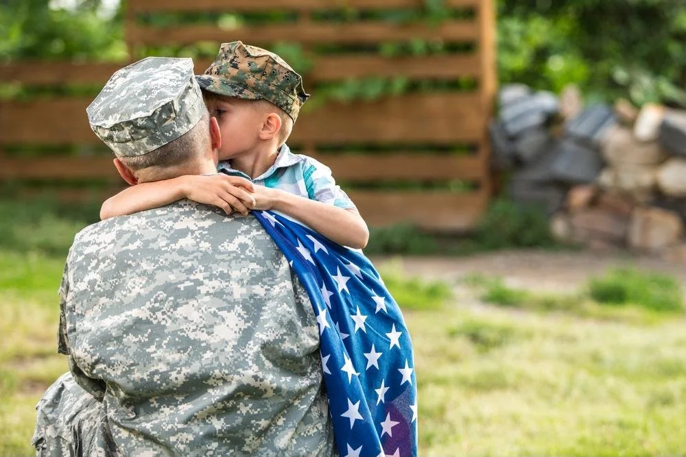 A soldier in camouflage uniform holding a young boy in military attire, who is kissing him, outdoors in a backyard setting.
