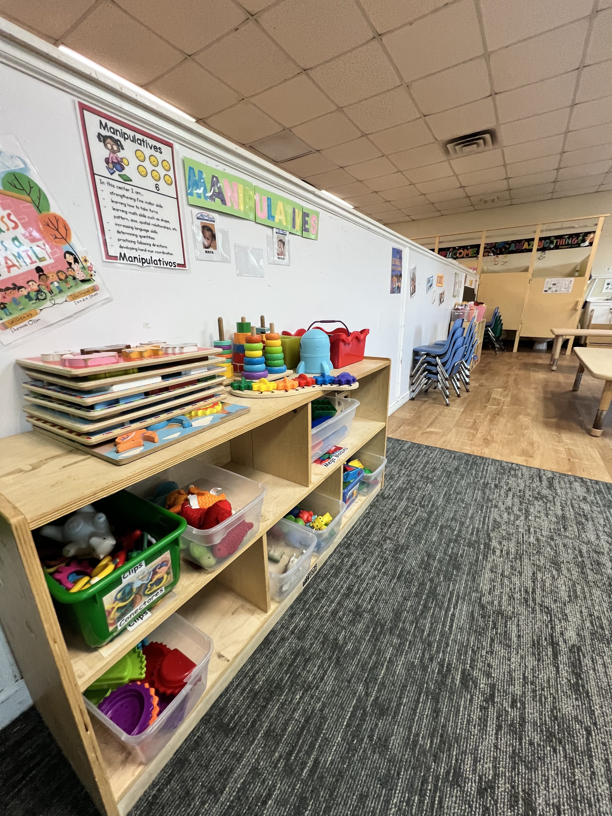 A classroom with a wooden shelf filled with various colorful toys and educational materials. The wall behind has educational posters, including one titled 'Manipulatives'. To the right, there are stacked chairs and tables, and a carpeted floor area.