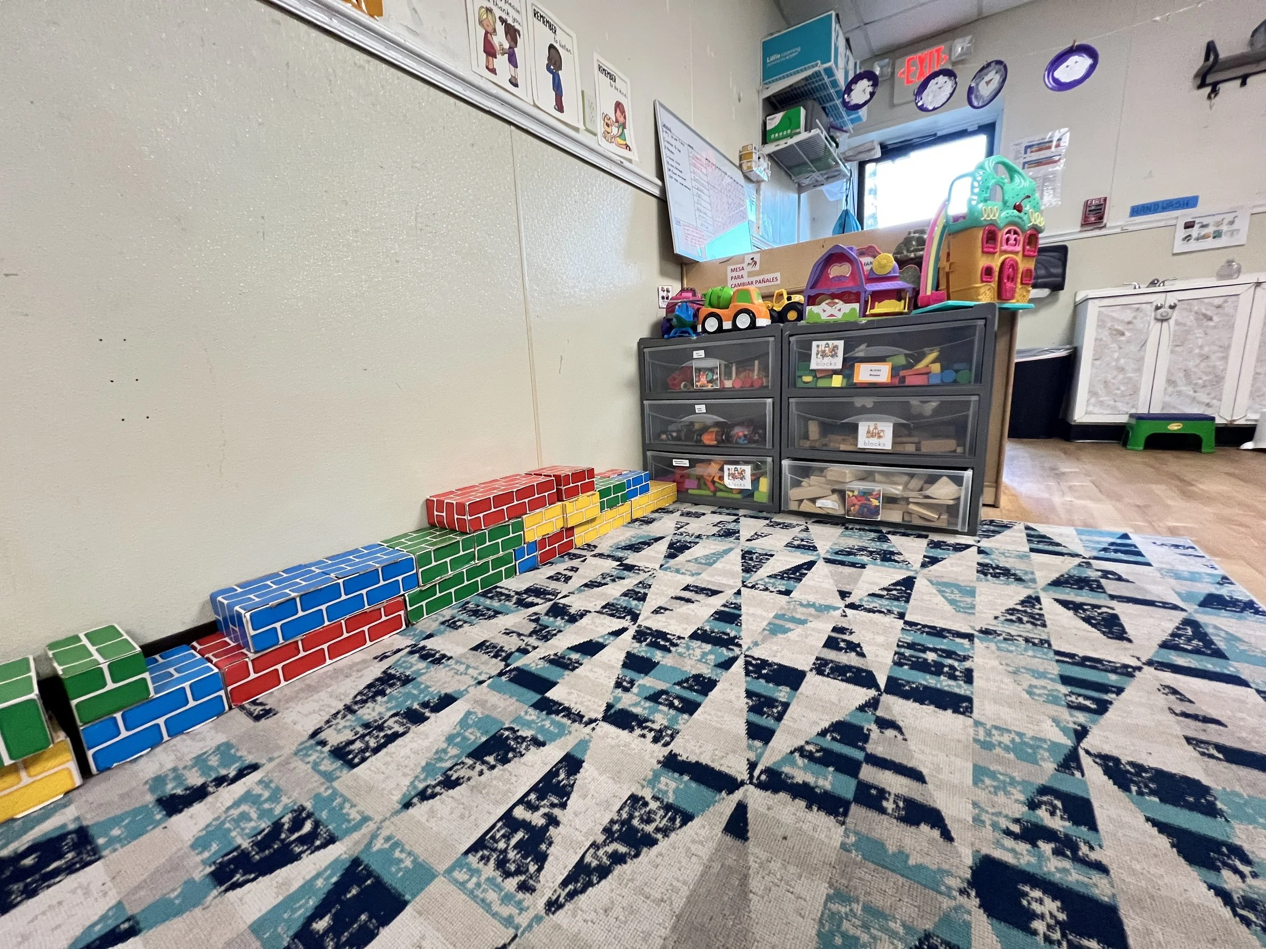 Colorful toy blocks are arranged in a staircase pattern on the floor near a plastic storage cabinet filled with various toys and blocks. The background includes posters on the wall, a window, and some hanging decorations.