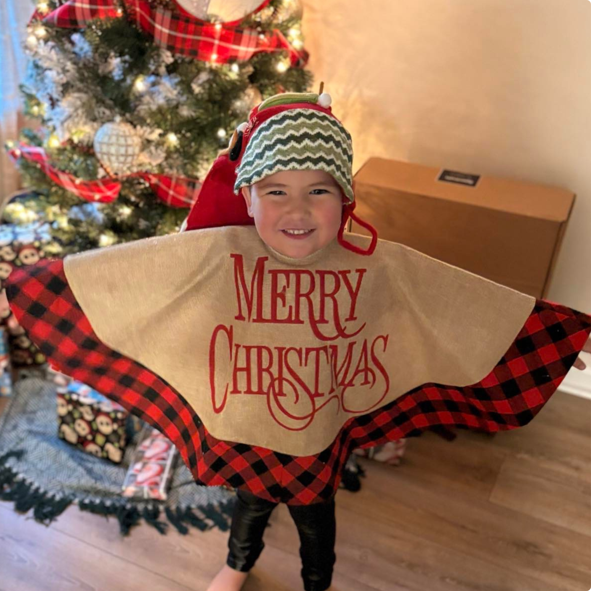 A young boy dressed in a Christmas-themed outfit, holding a gift bag with 'Merry Christmas' printed on it, in front of a decorated Christmas tree with ornaments and lights.