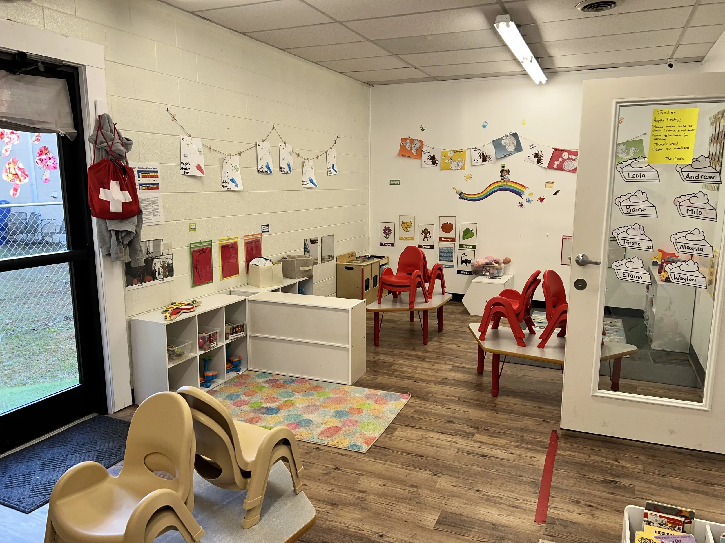 A colorful classroom with educational wall decorations, children’s chairs, and toys. There's a rainbow wall art, educational posters, and a whiteboard with children's names and drawings. A window with a view outside and a door are visible.