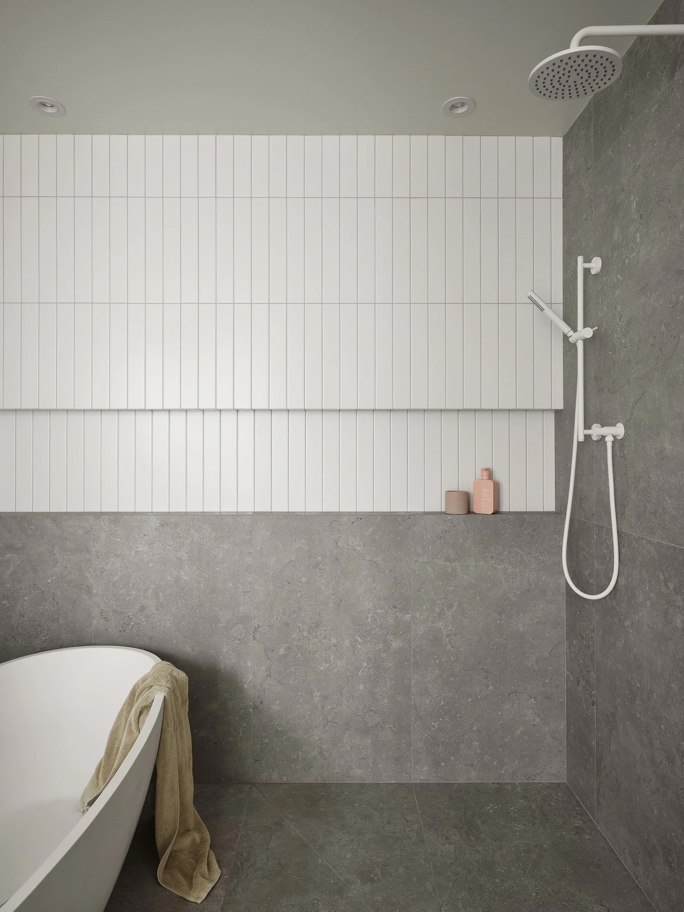Salle de bain moderne avec douche à l'italienne, mur en carrelage blanc, sol en pierre grise, baignoire blanche, serviette beige, bouteille de savon rose, pommeau de douche blanc.