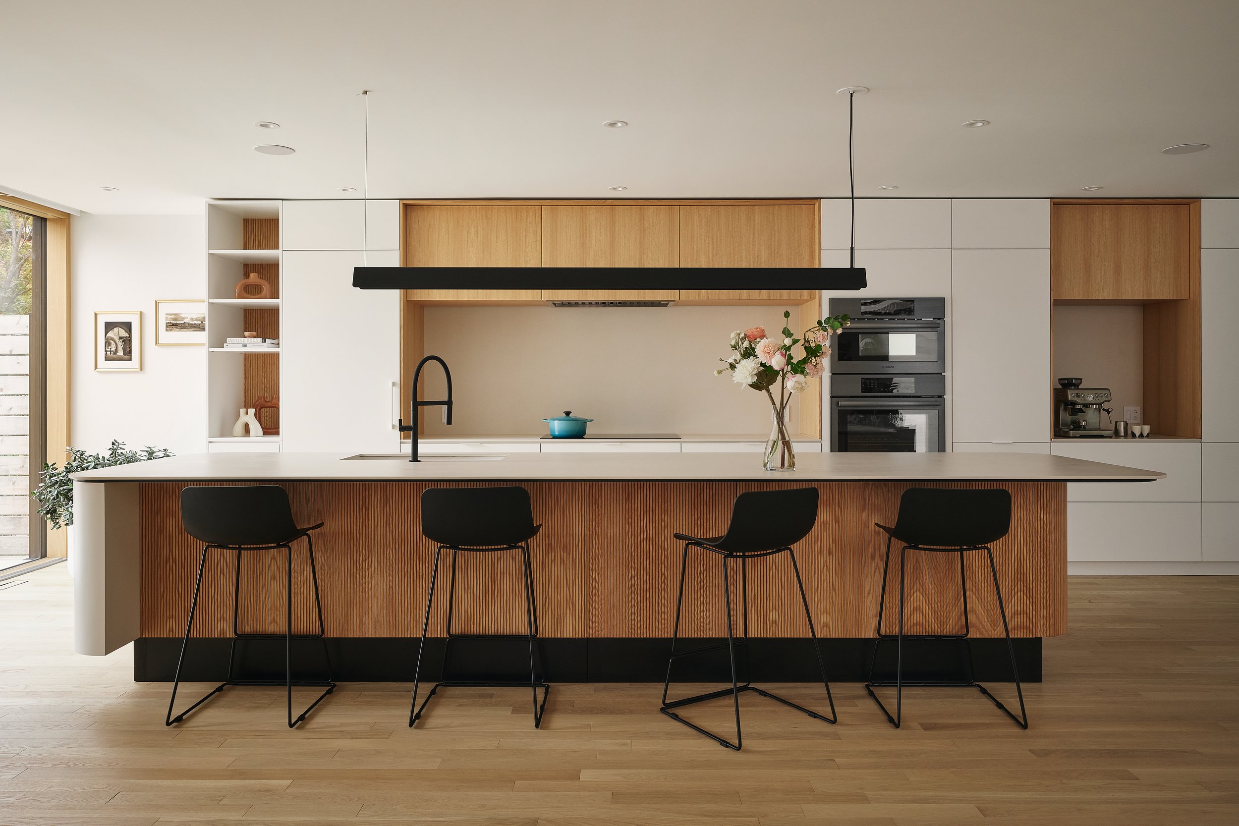 Une cuisine moderne avec un îlot central en bois clair, des chaises noires en métal, un vase avec des fleurs, un four encastré, un robinet noir, et un peu de décoration simple