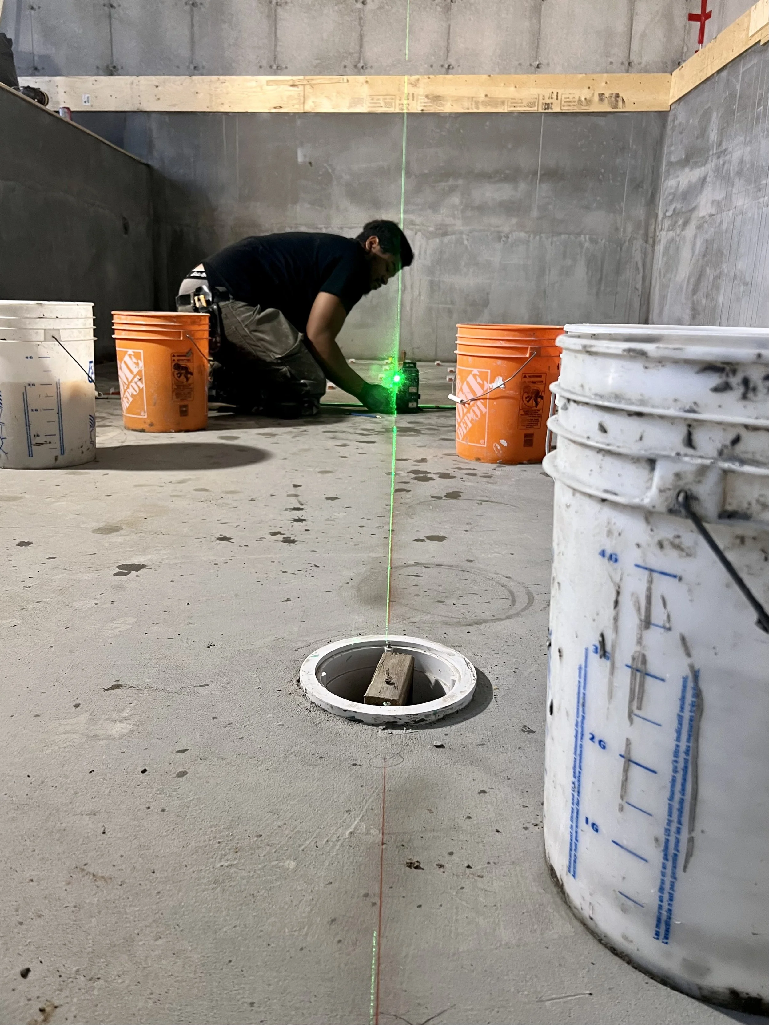 Un homme en tenue de construction utilisant un instrument laser vert pour prendre des mesures ou des alignements sur un chantier de construction. Il est entouré de buckets en plastique blancs et orange, placés sur un sol en béton. La scène se déroule