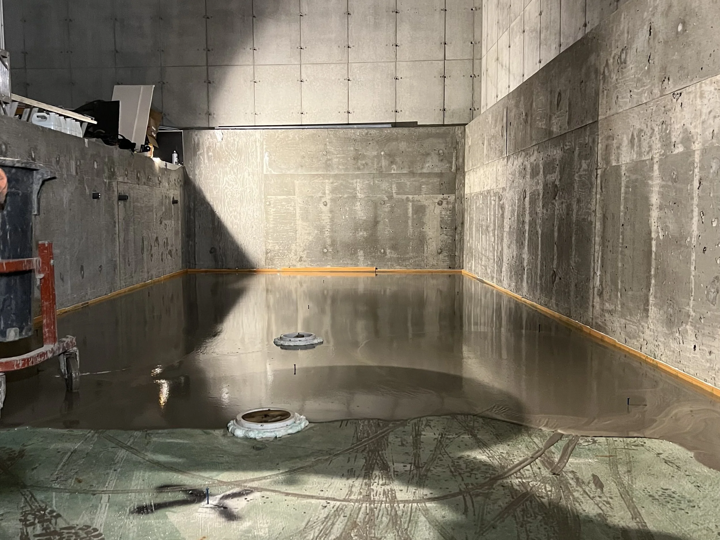 Un espace intérieur en construction avec des murs en béton brut, un sol en béton poli, et des tuyaux et équipements en cours d'installation.