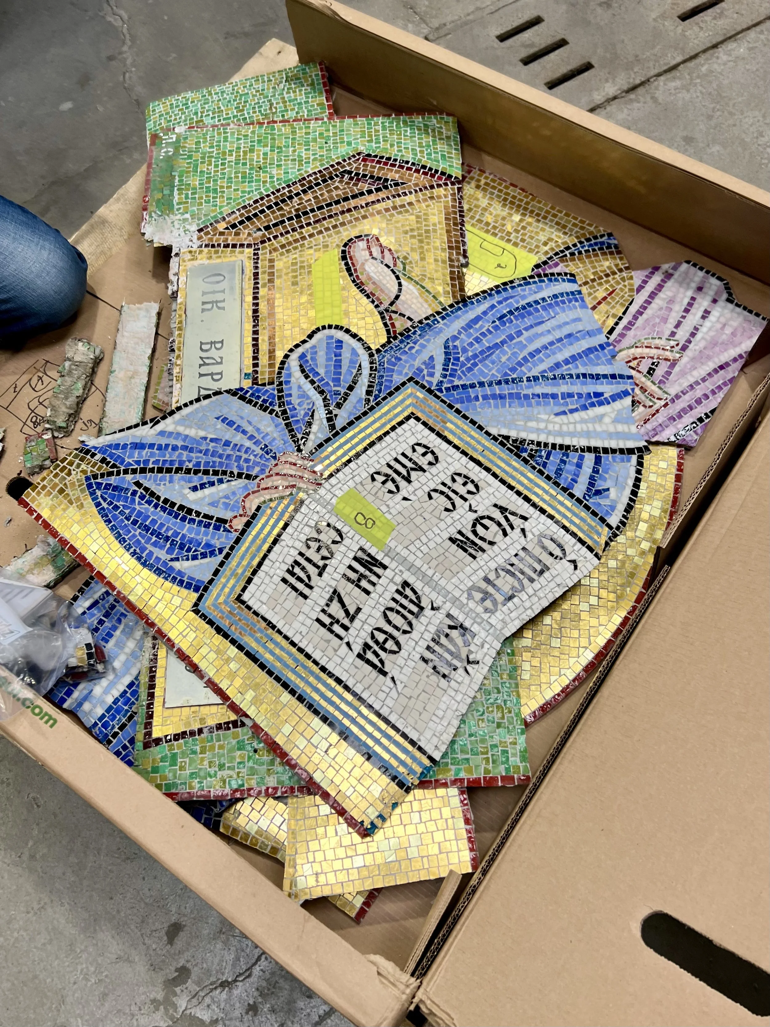 Des morceaux de mosaïque colorés en vert, marron, jaune, bleu et rose, représentant une image de livre ou de panneau avec des mots en français, posés dans une boîte en carton sur un sol en béton.