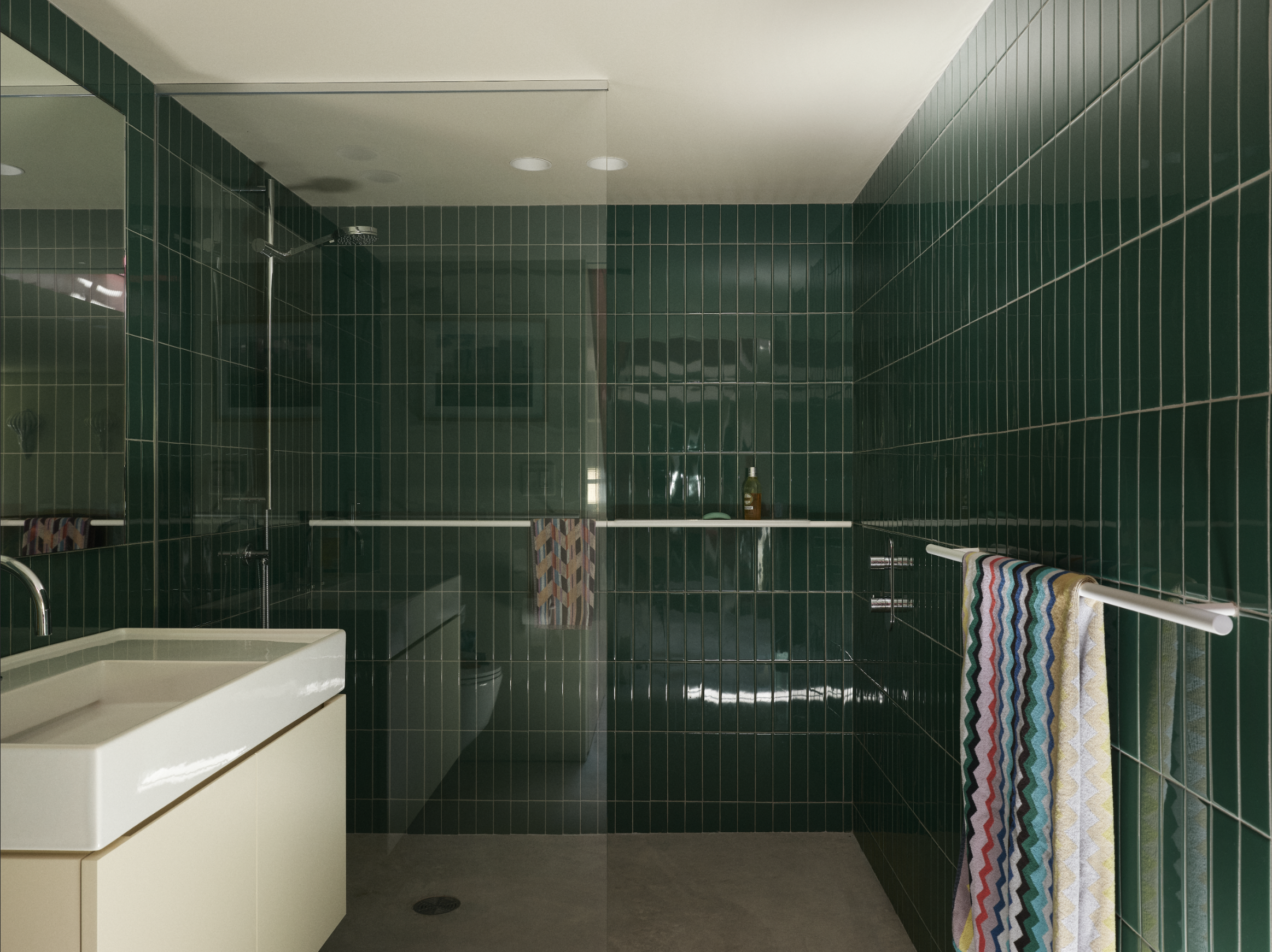 Salle de bain avec murs en carrelage vert, douche à l'italienne, lavabo blanc avec meuble, serviettes colorées et miroir au plafond.