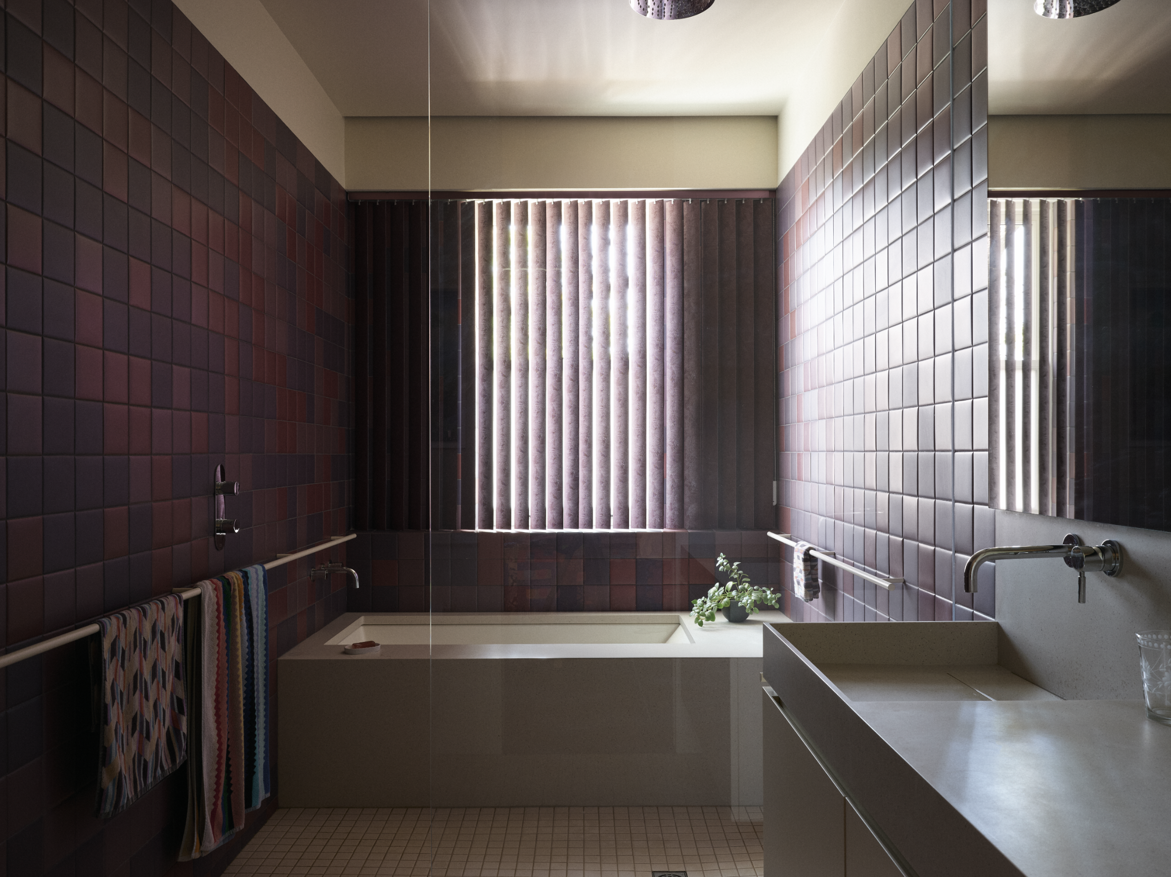 Salle de bain moderne avec des carreaux de couleur bordeaux, une fenêtre avec des stores, un lavabo blanc avec des plantes et des serviettes colorées sur une barre.