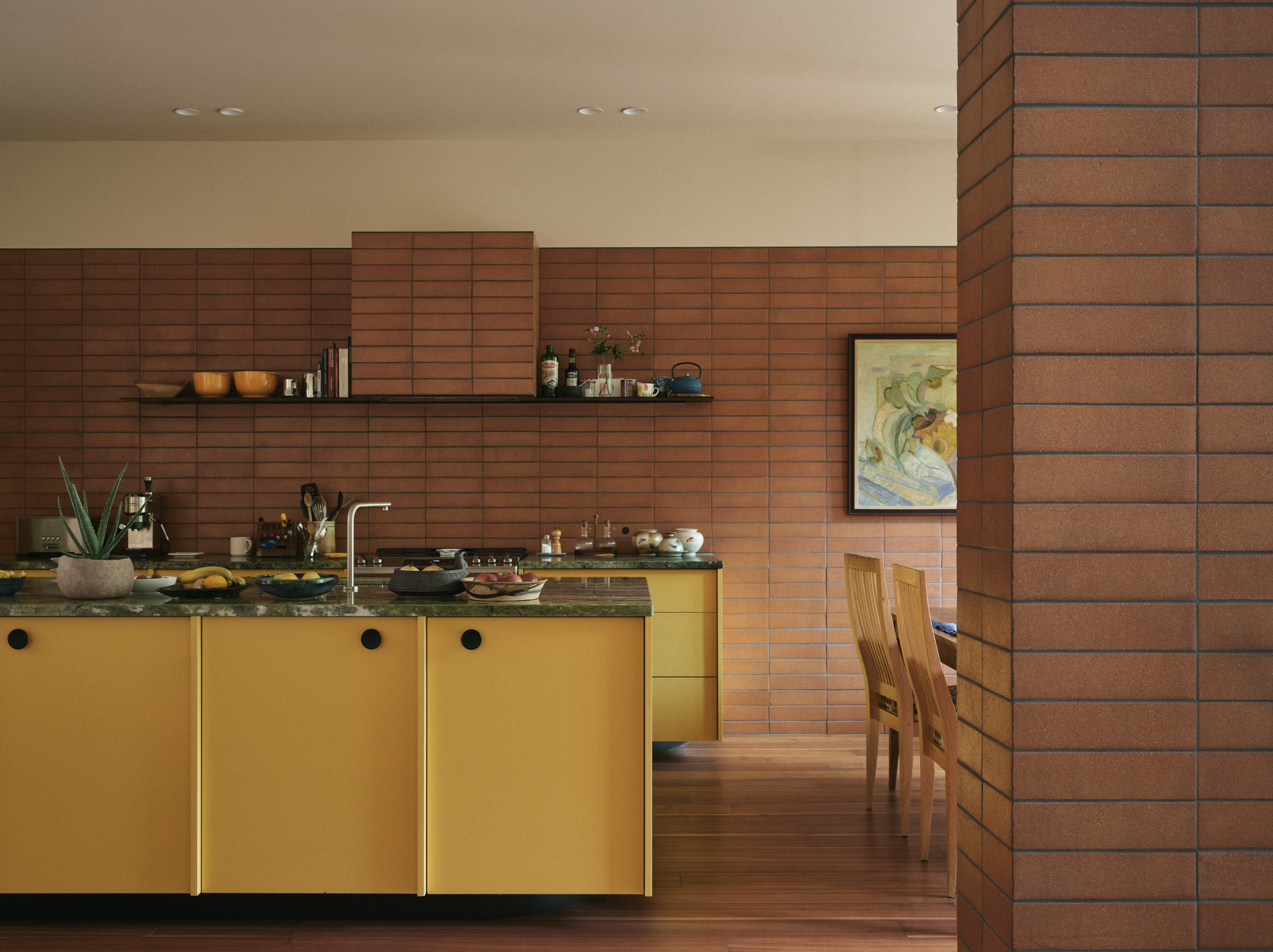 Cuisine moderne avec un comptoir jaune, murs en briques rouges, étagère avec des livres et objets, plantes, affiches et un coin repas avec chaises en bois.