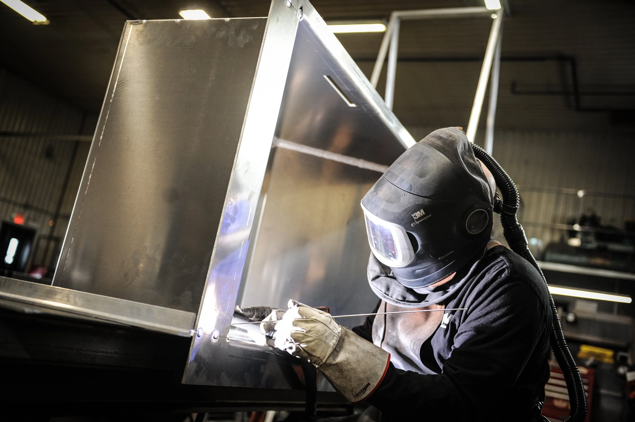 Un personne portant un casque de soudure et des gants de protection effectue une soudure sur une structure métallique dans un atelier industriel.