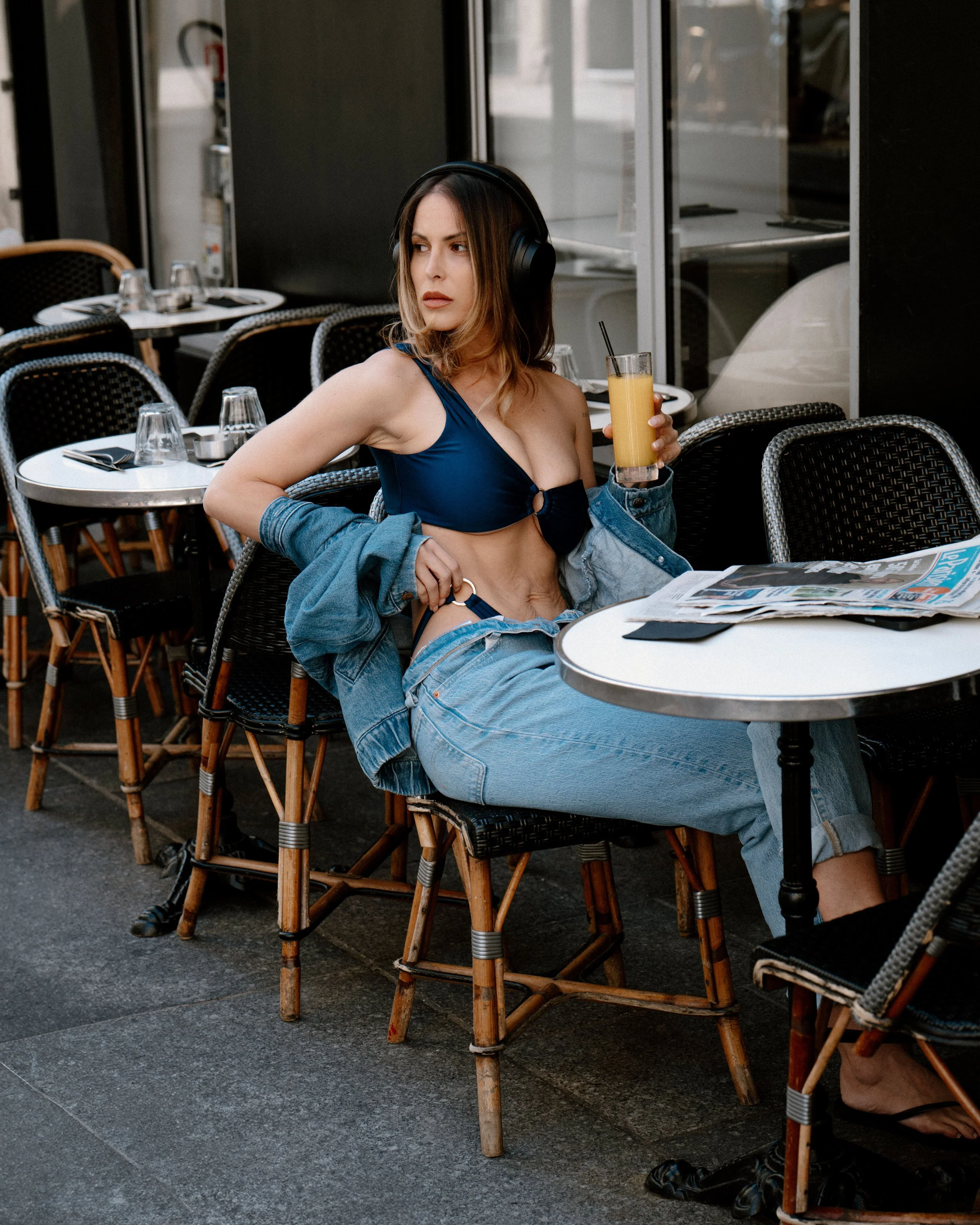 Une femme assise dans un restaurant ou un café, portant un soutien-gorge bleu, un jean décontracté et des écouteurs. Elle tient un verre de jus d'orange dans sa main. La table a une nappe blanche avec des verres, des fourchettes et un journal.