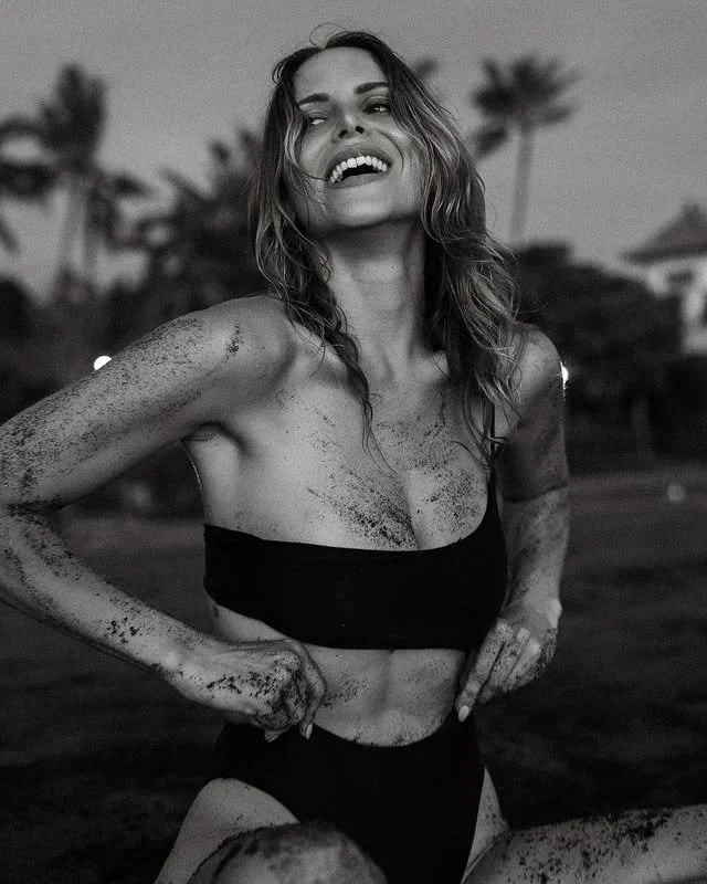 Femme souriante portant un haut noir et un pantalon, avec de la terre sur le corps, en plein air, palmier en arrière-plan, image en noir et blanc.