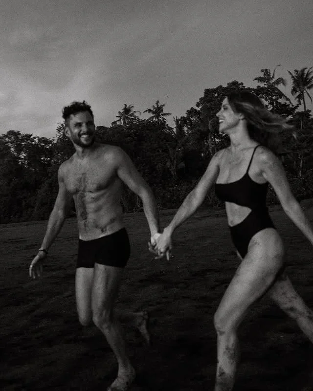 Un couple souriant, habillé en maillots de bain, court main dans la main sur une plage avec des palmiers en arrière-plan, dans un décor tropical