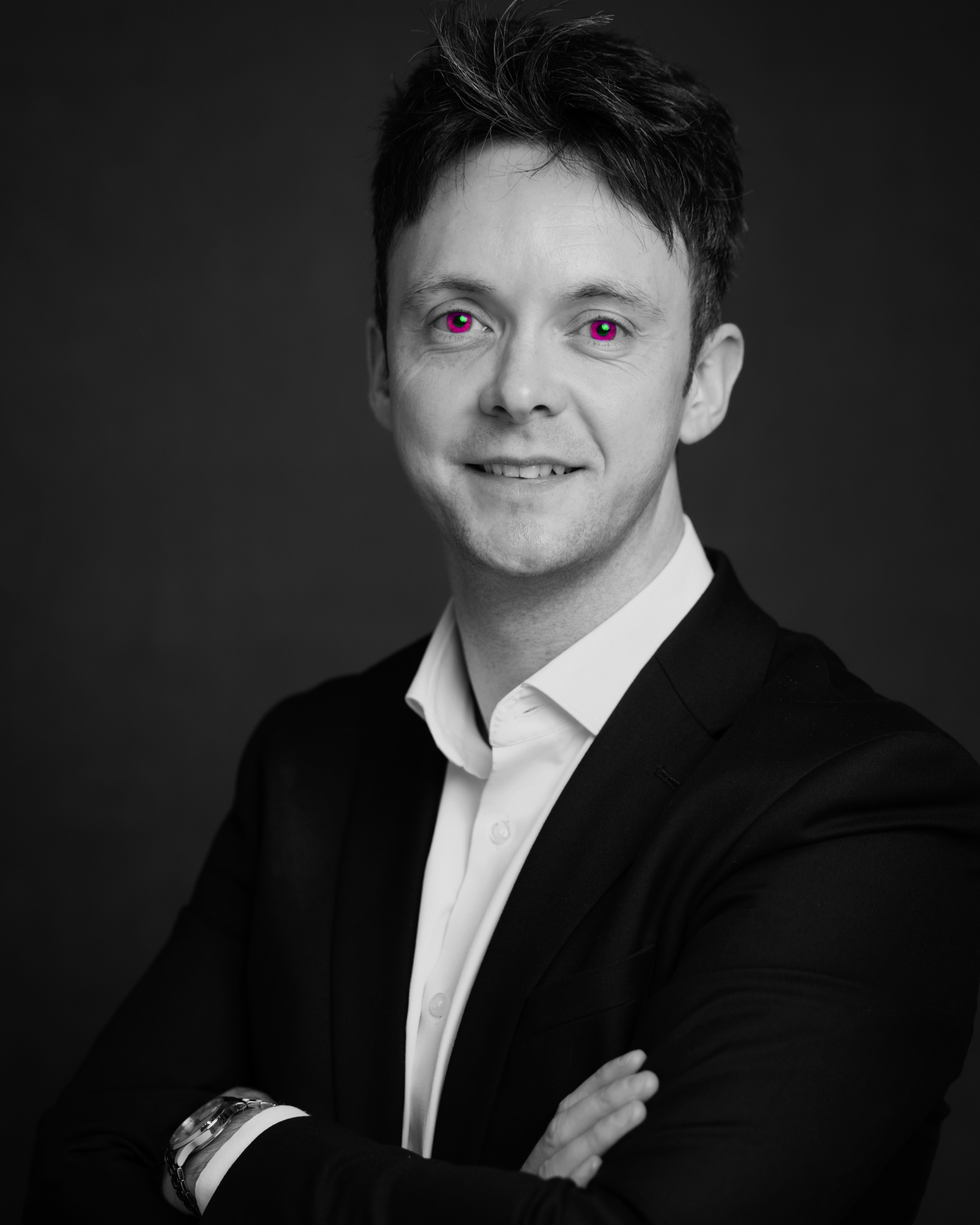 Black and white portrait of a Daniel Ring, CEO of Wicked Flamingo wearing a dark suit with a white shirt, crossed arms, and a watch on his wrist, against a dark background.