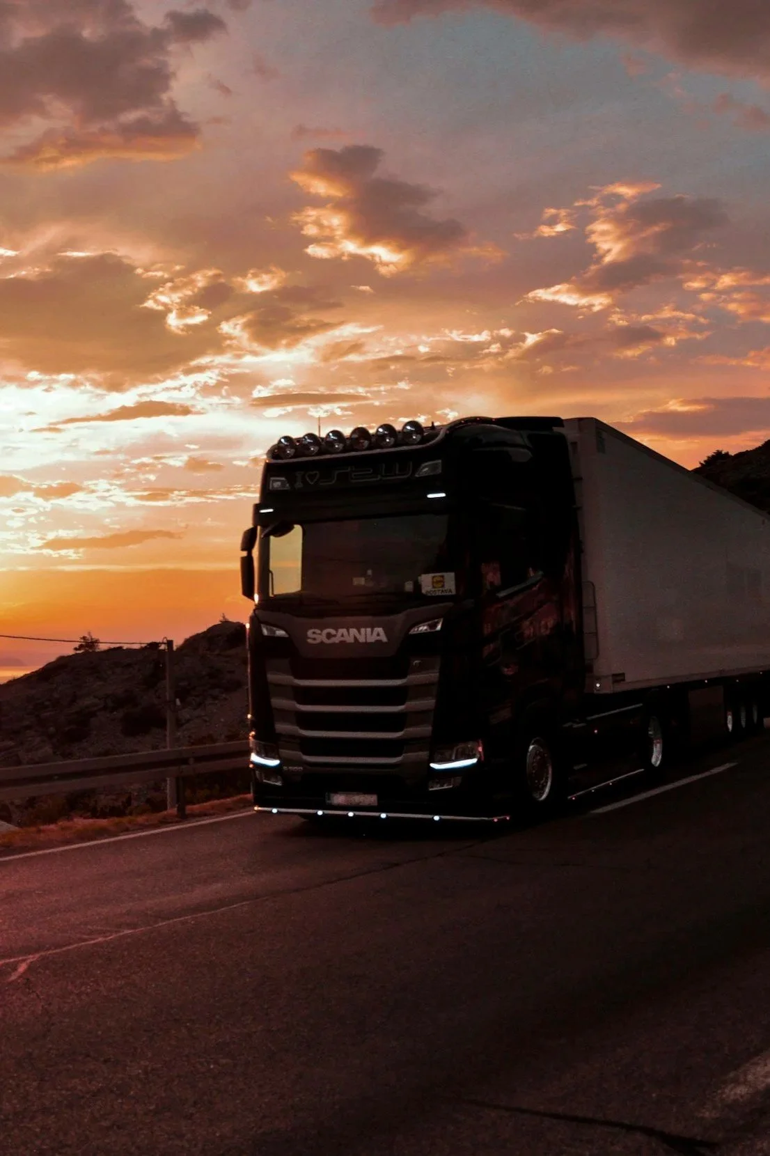 A black semi-truck traveling on a highway at sunset with a colorful sky and clouds in the background.