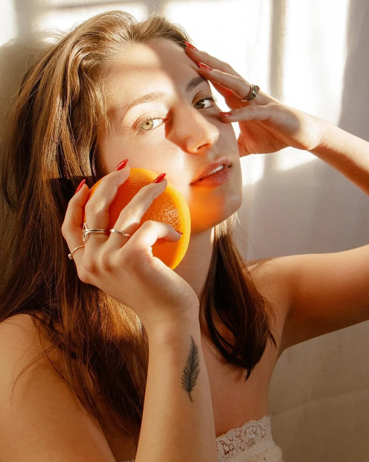 A woman with red hair and light skin holding a grapefruit near her face, touching her forehead with one hand, with sunlight illuminating her face against a light background.