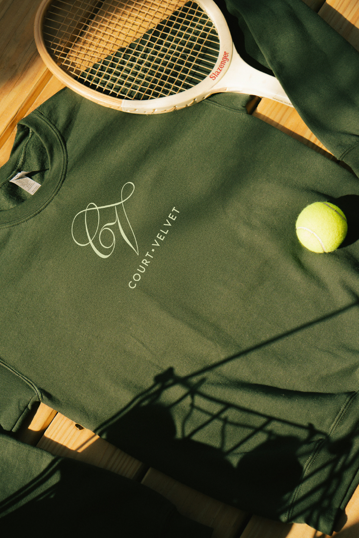 A green sweatshirt with the words 'COURT VELVET' and a logo, a yellow tennis ball, a tennis racket, and a wooden surface with shadows.