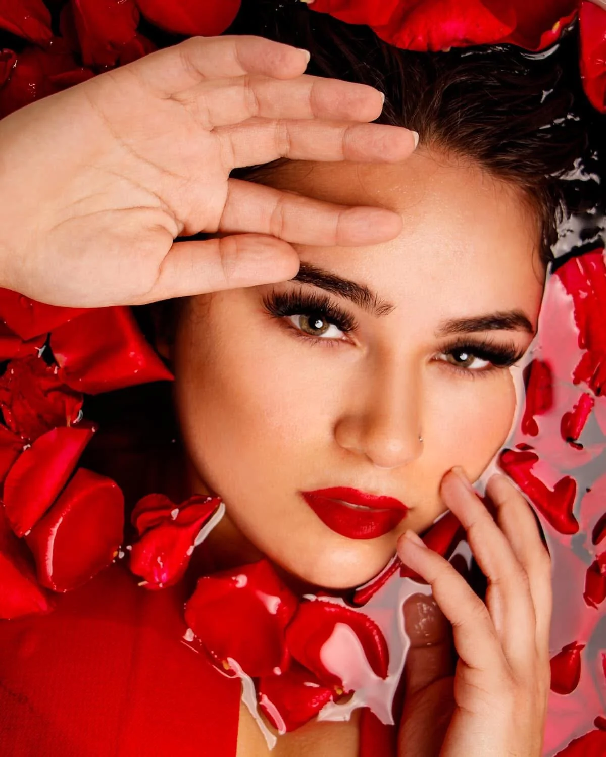 A woman with dark hair and makeup, including red lipstick and false eyelashes, floats in water surrounded by red rose petals, holding her hand near her face, with a confident expression.