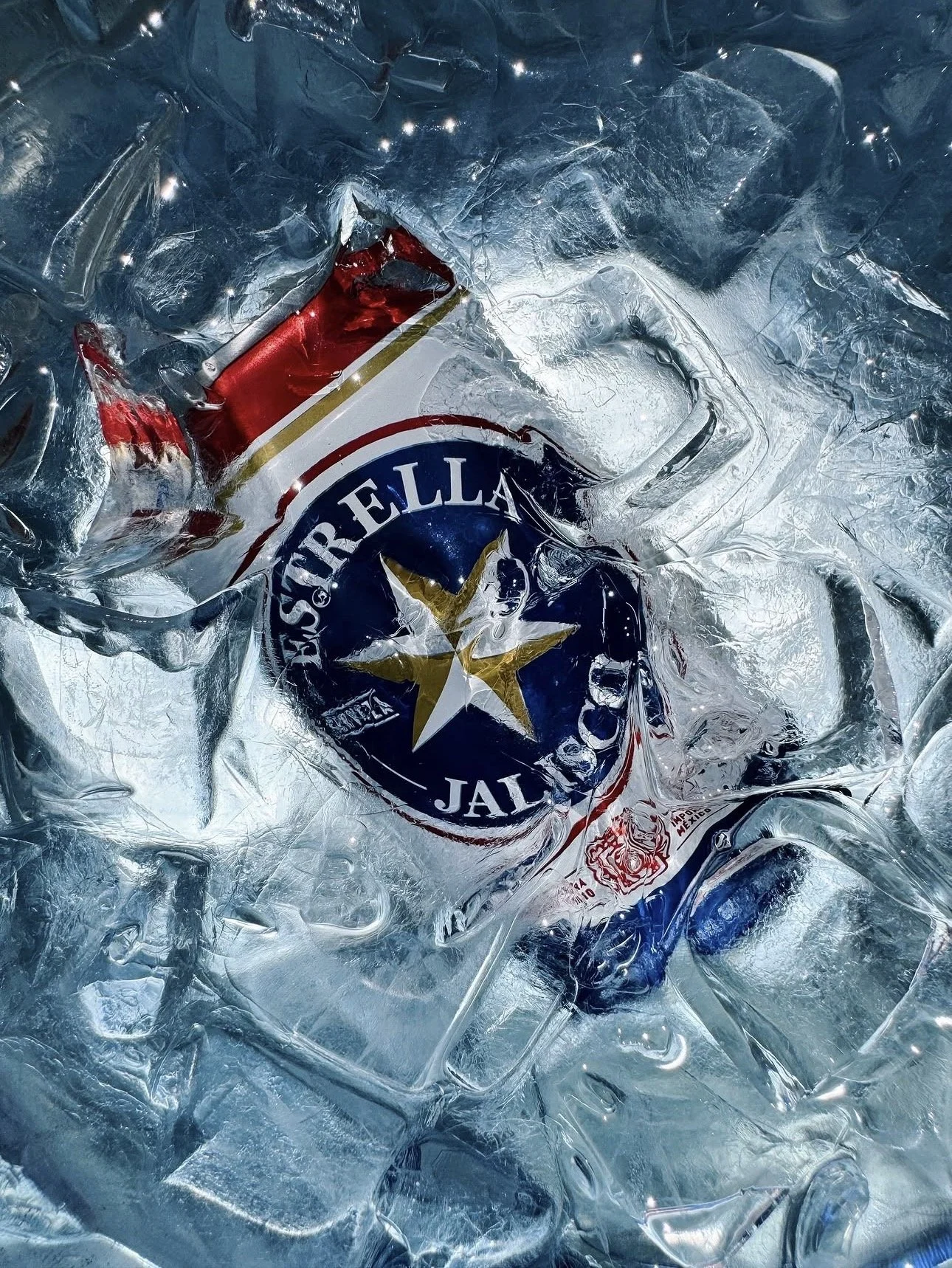 Crushed ice surrounding a partially melted can of 'Cerveza Modelo' beer with the logo visible, the can is red and white with a star, amidst reflective ice and condensation.
