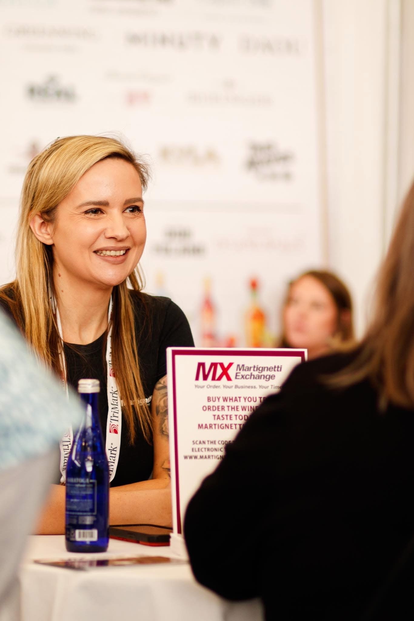 A woman smiling at a booth with a sign that reads 'MIX Martignetti Exchange' at a conference or trade show.