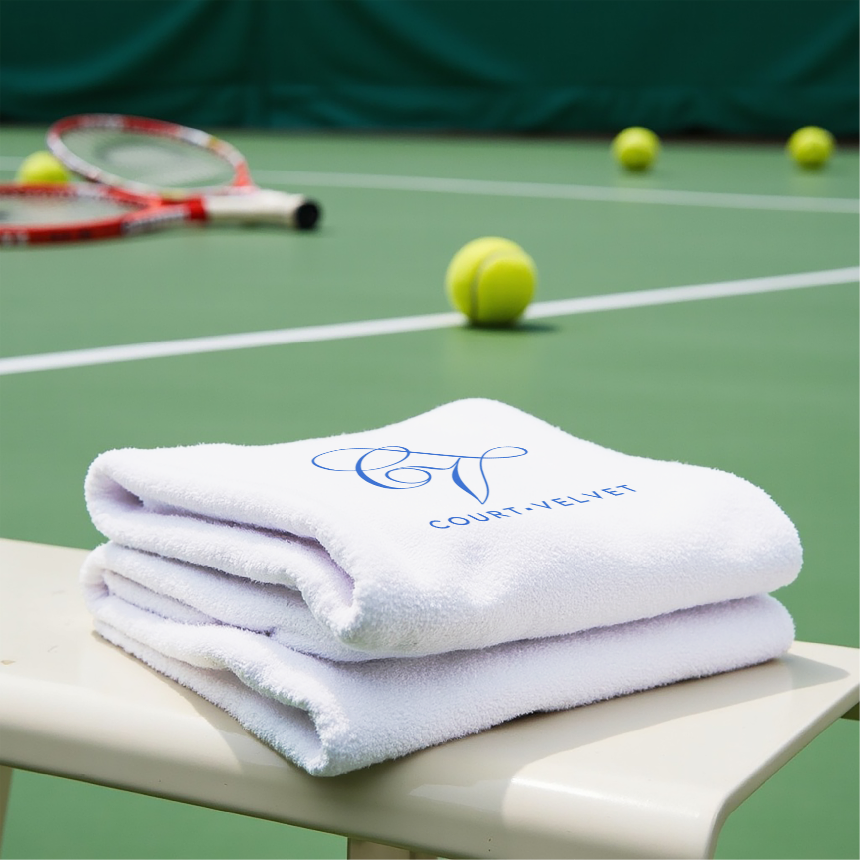 Stack of three folded white towels with a blue embroidered logo, tennis rackets and tennis balls on a green tennis court.