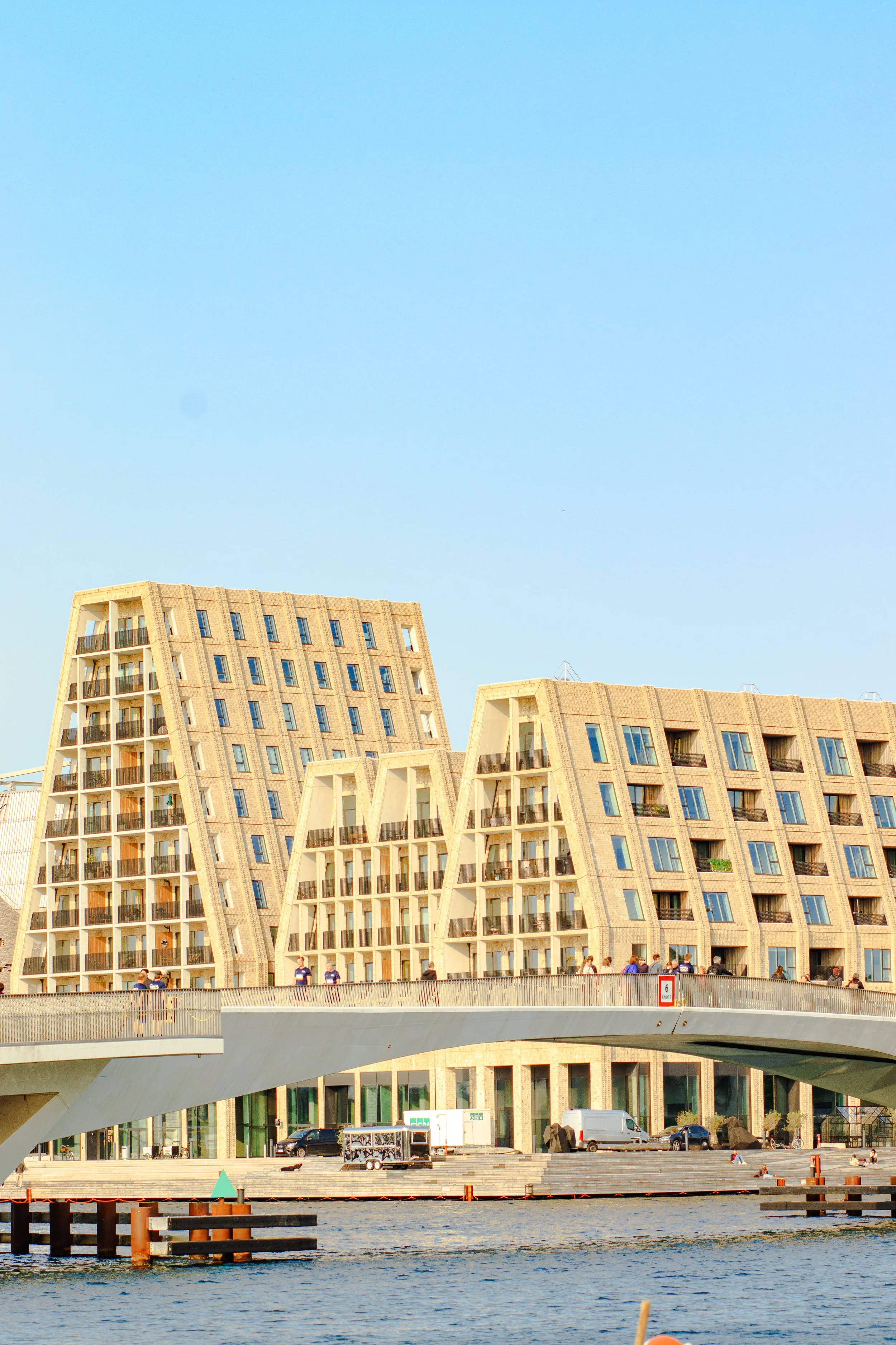 Modern, multi-story residential buildings with a distinctive angular architectural design, located by a waterfront. The buildings have large windows and balconies, and the lower level includes commercial spaces. There is a bridge with pedestrians on 