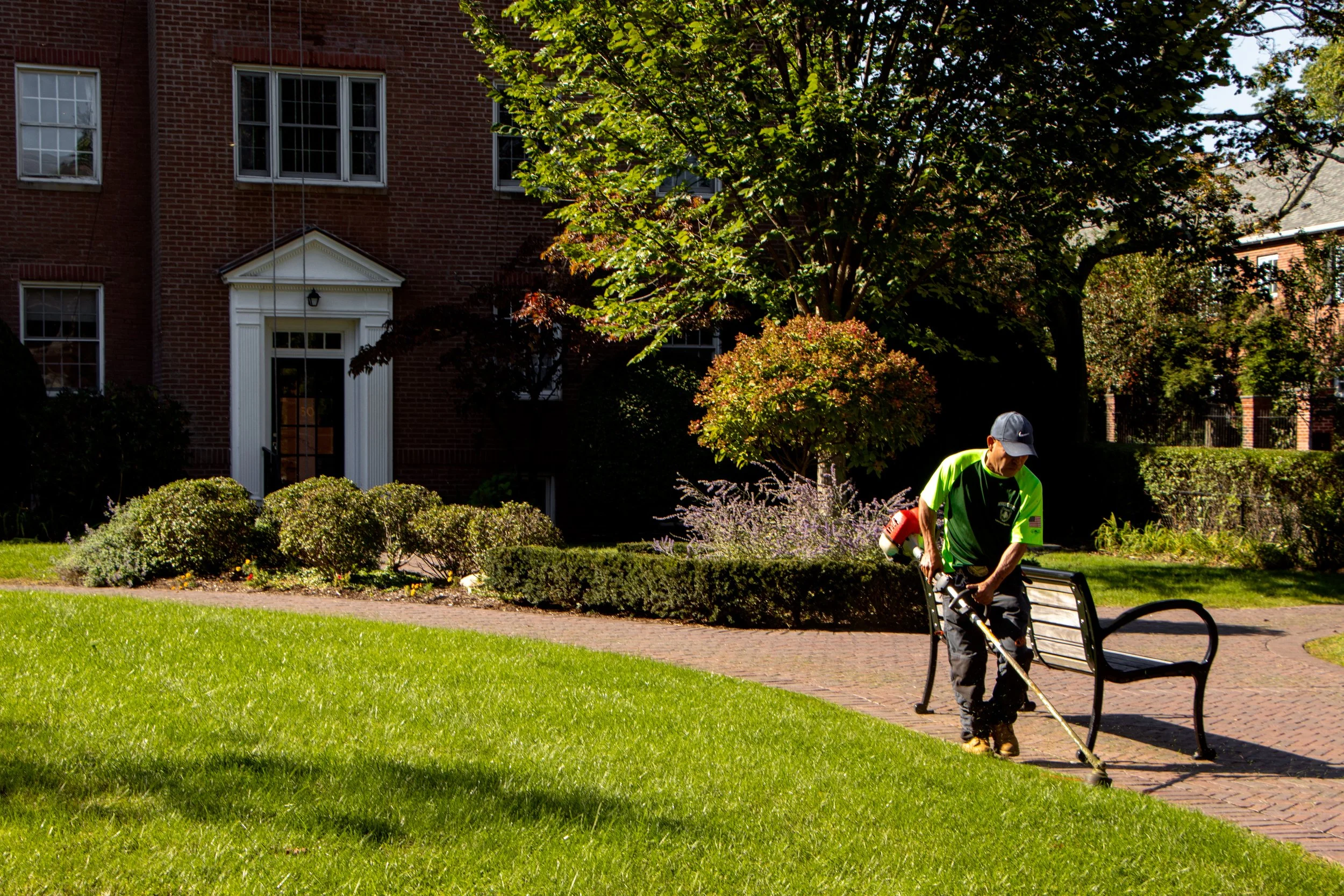 A man in a neon green shirt and dark cap trimming bushes in a front yard with a weed trimmer. In the background, there's a brick house with white-framed windows, a white front door with a porch roof, and a large tree with green leaves. The yard has g