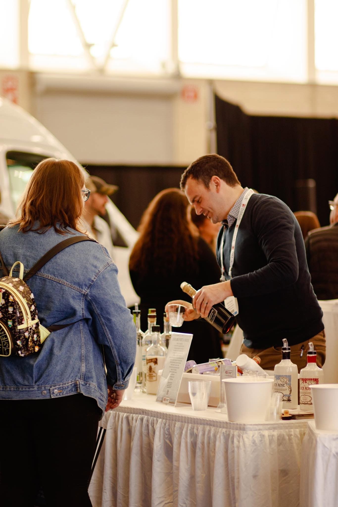 A man pouring a drink at a booth at a trade show or convention, with several bottles and informational materials on the table, and a woman with a backpack standing nearby.