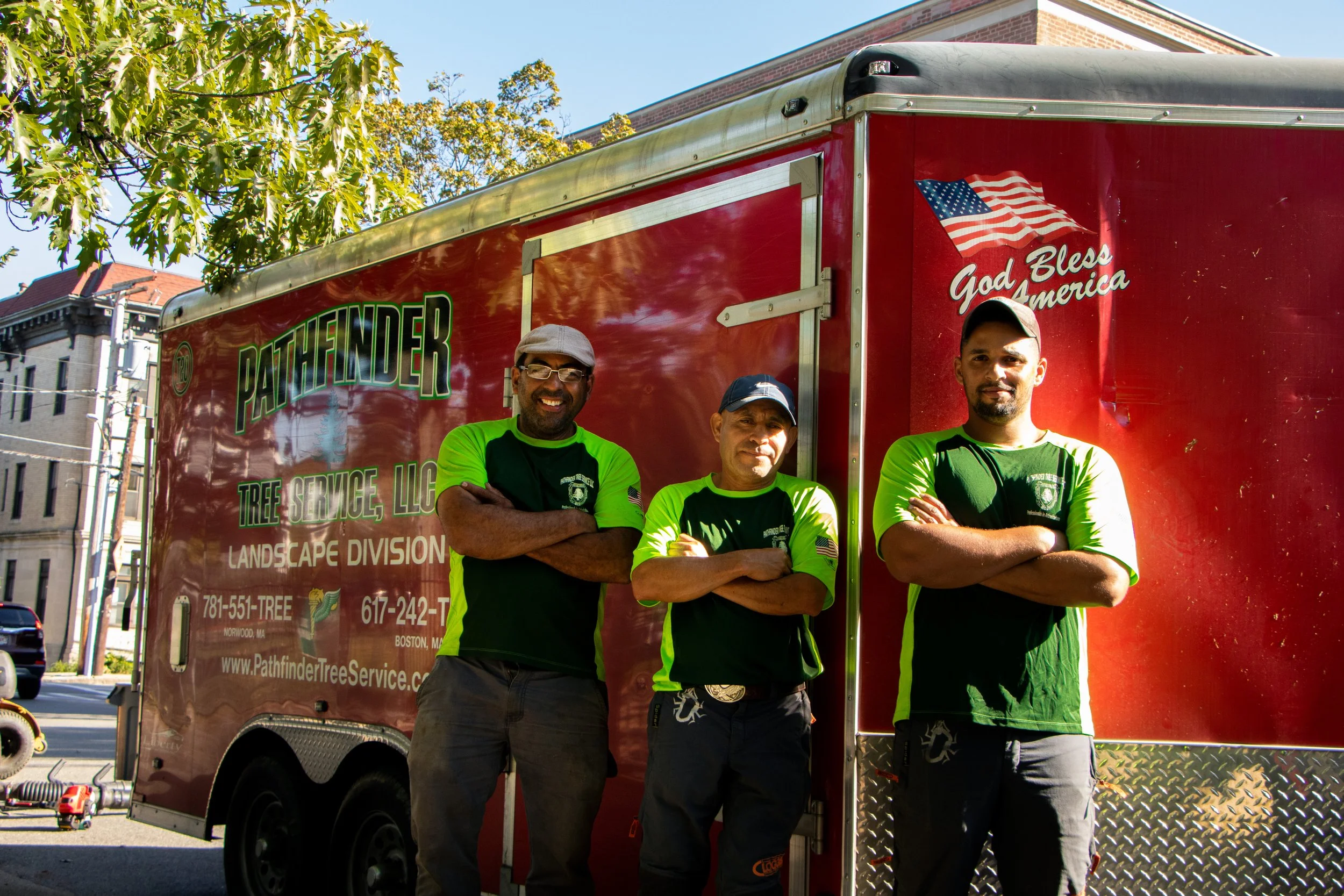 Three men standing with arms crossed in front of a red tree service truck that has an American flag decal and the words "God Bless America" on it. The men are wearing bright green and black work uniforms and are standing outdoors on a city street wit
