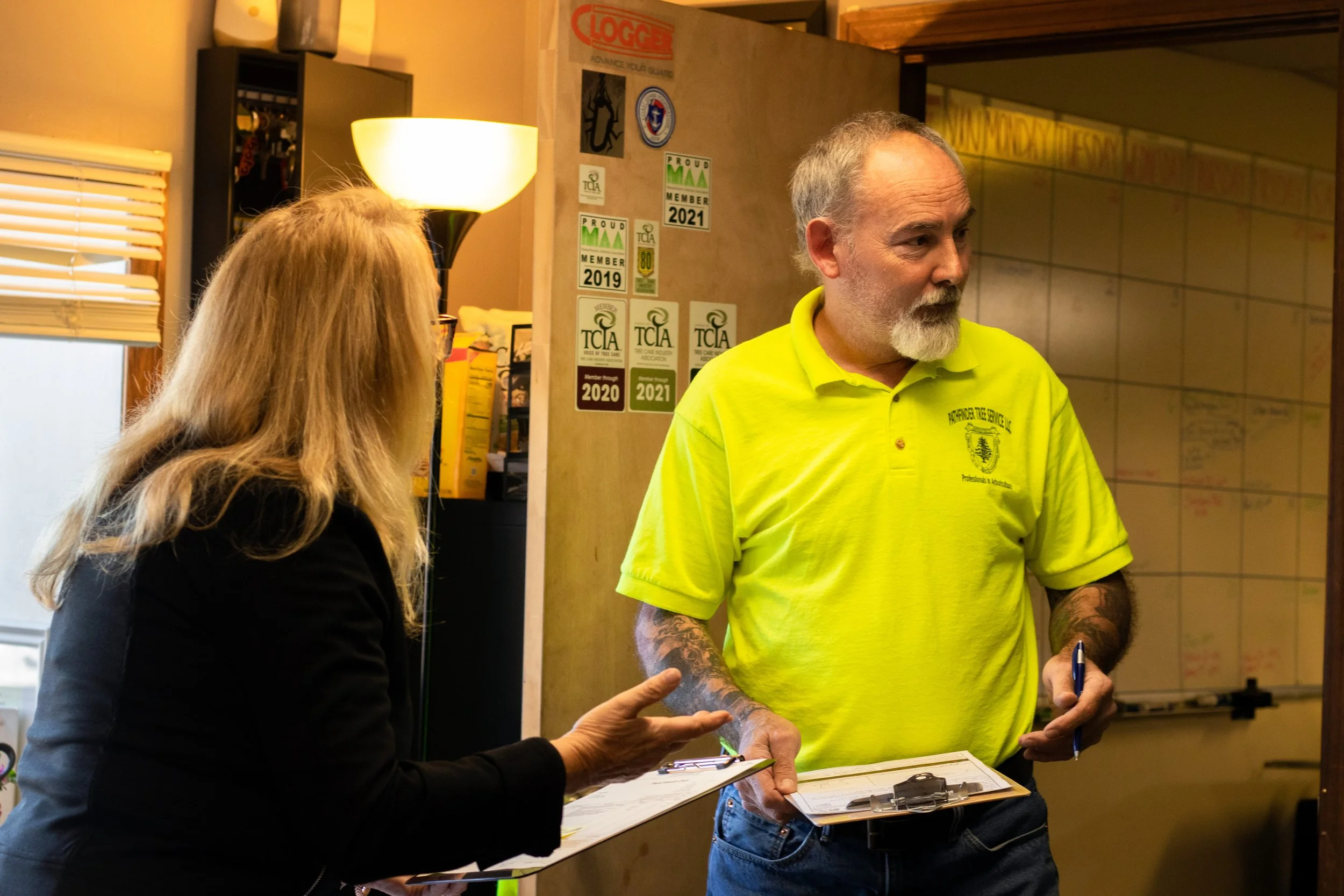 A man with gray hair and a beard wearing a bright yellow shirt standing indoors, engaging in conversation with a woman with long blonde hair who is holding a clipboard. The background shows various stickers and posters on the wall, a window with blin