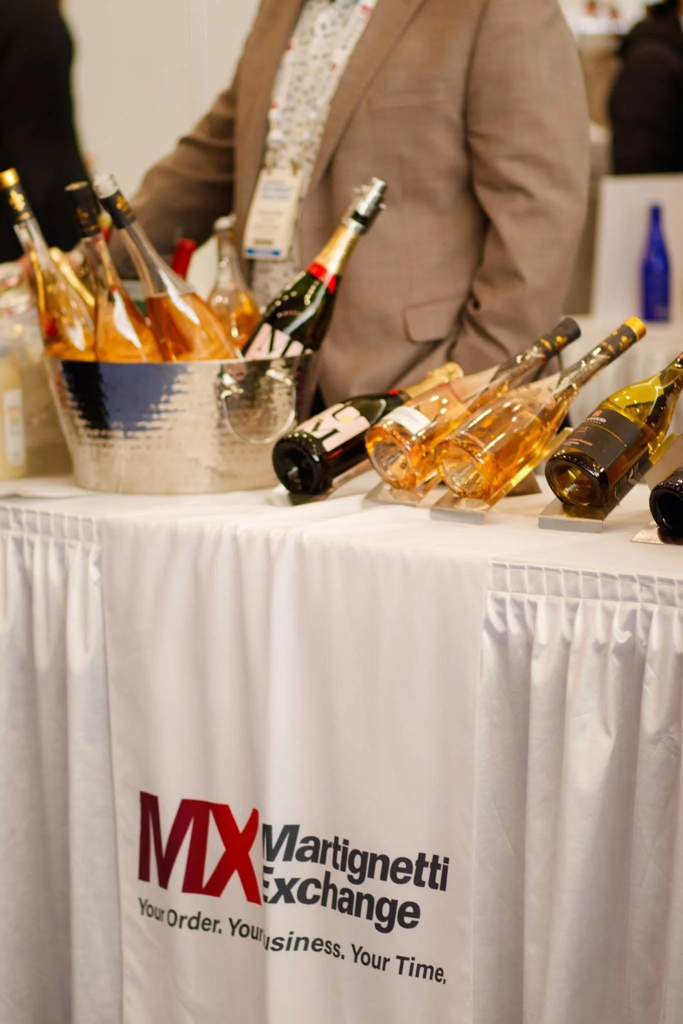 Table displaying various bottles of wine or champagne at an event or trade show, with a person standing behind the table. The tablecloth has a logo with the text "MX Martignetti Exchange" and a tagline beneath.