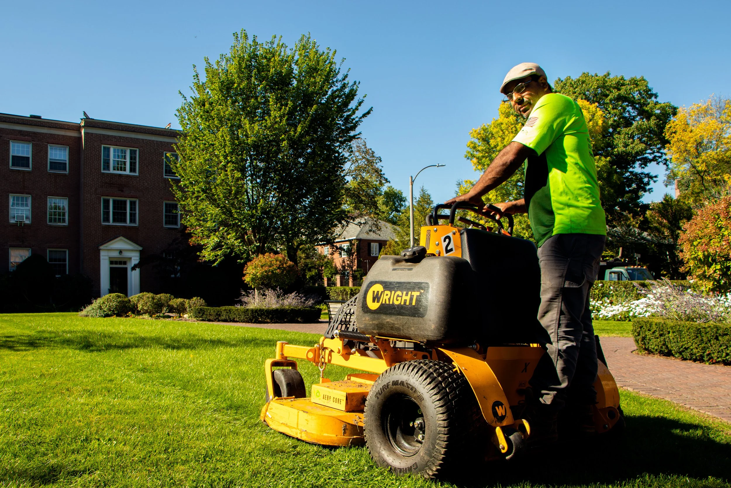 A man operating a yellow Wright brand lawn mower on a green lawn, with trees and a brick building in the background, under a clear blue sky.